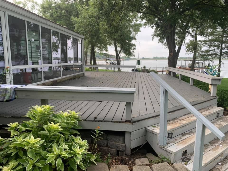 A grey wooden deck attached to a screened porch, featuring built-in benches, stairs, and a view of a lake with trees.