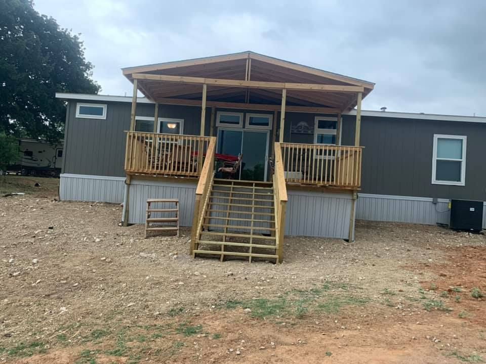 A grey mobile home with a wooden porch and stairs set on a dirt and gravel lot under a cloudy sky.