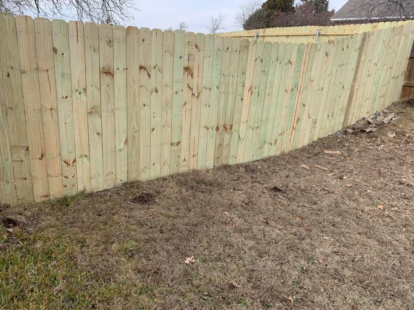 A new light-colored wood privacy fence stands in a yard with dry grass under an overcast sky.