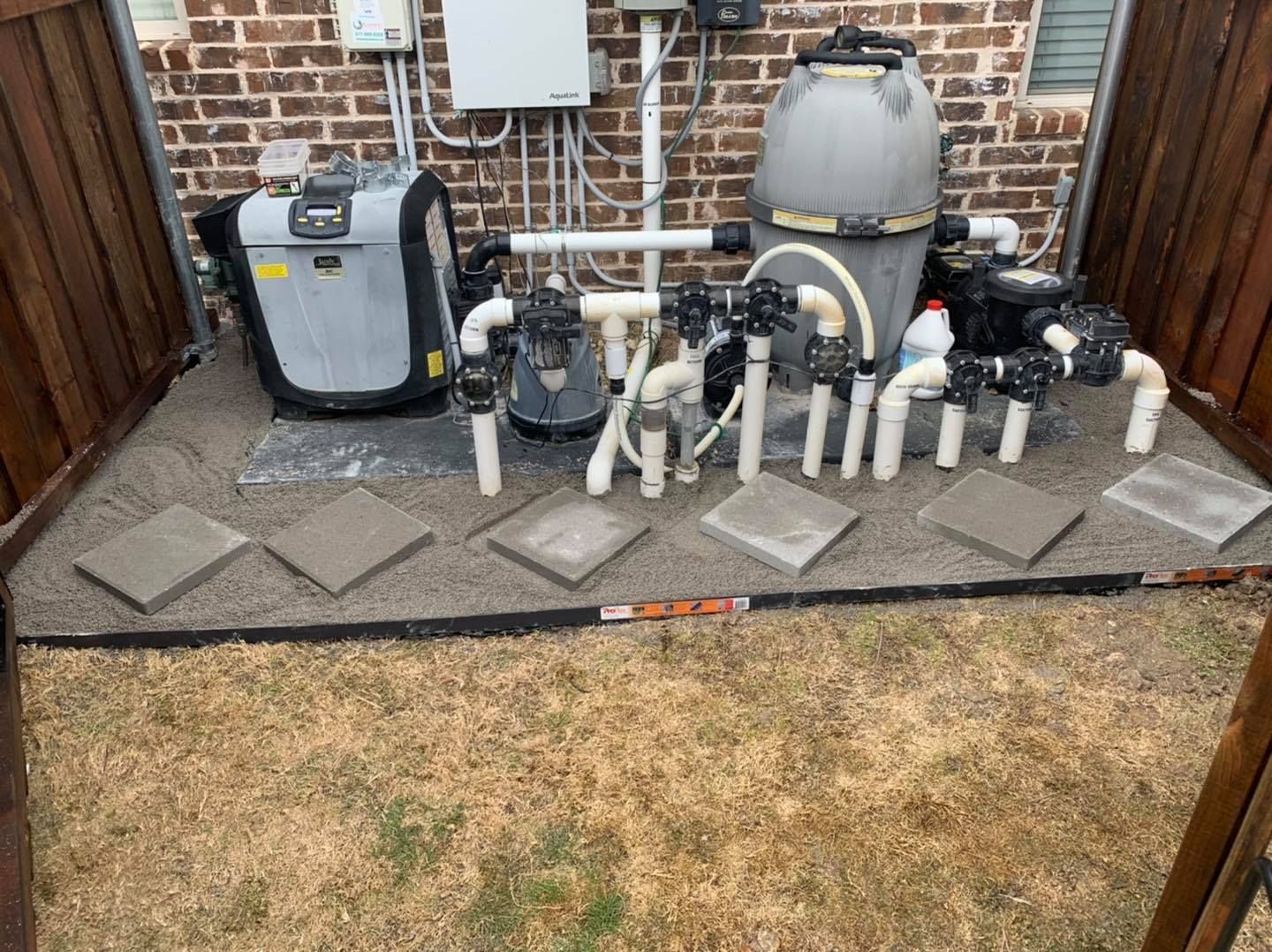 A pool equipment setup with a grey heater and filter tank, connected by white PVC pipes on a gravel pad near a brick wall.