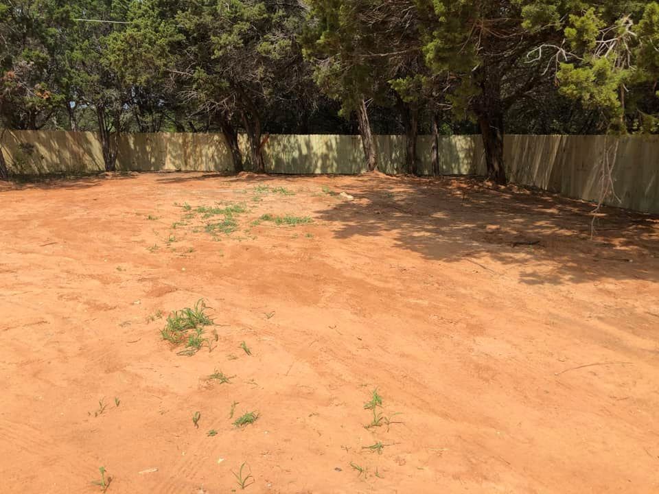A wide-angle shot of a bare, red dirt lot enclosed by a wooden fence with dense green trees in the background.