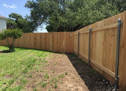 A wooden privacy fence with metal posts stands in a grassy backyard under a clear blue sky.