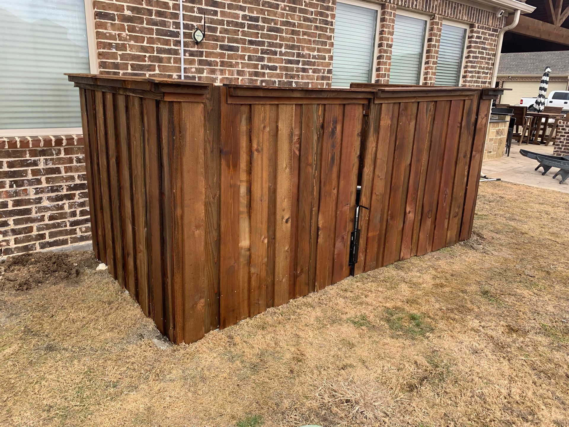A stained wooden privacy fence screen stands against a brick house exterior on a sunny day.