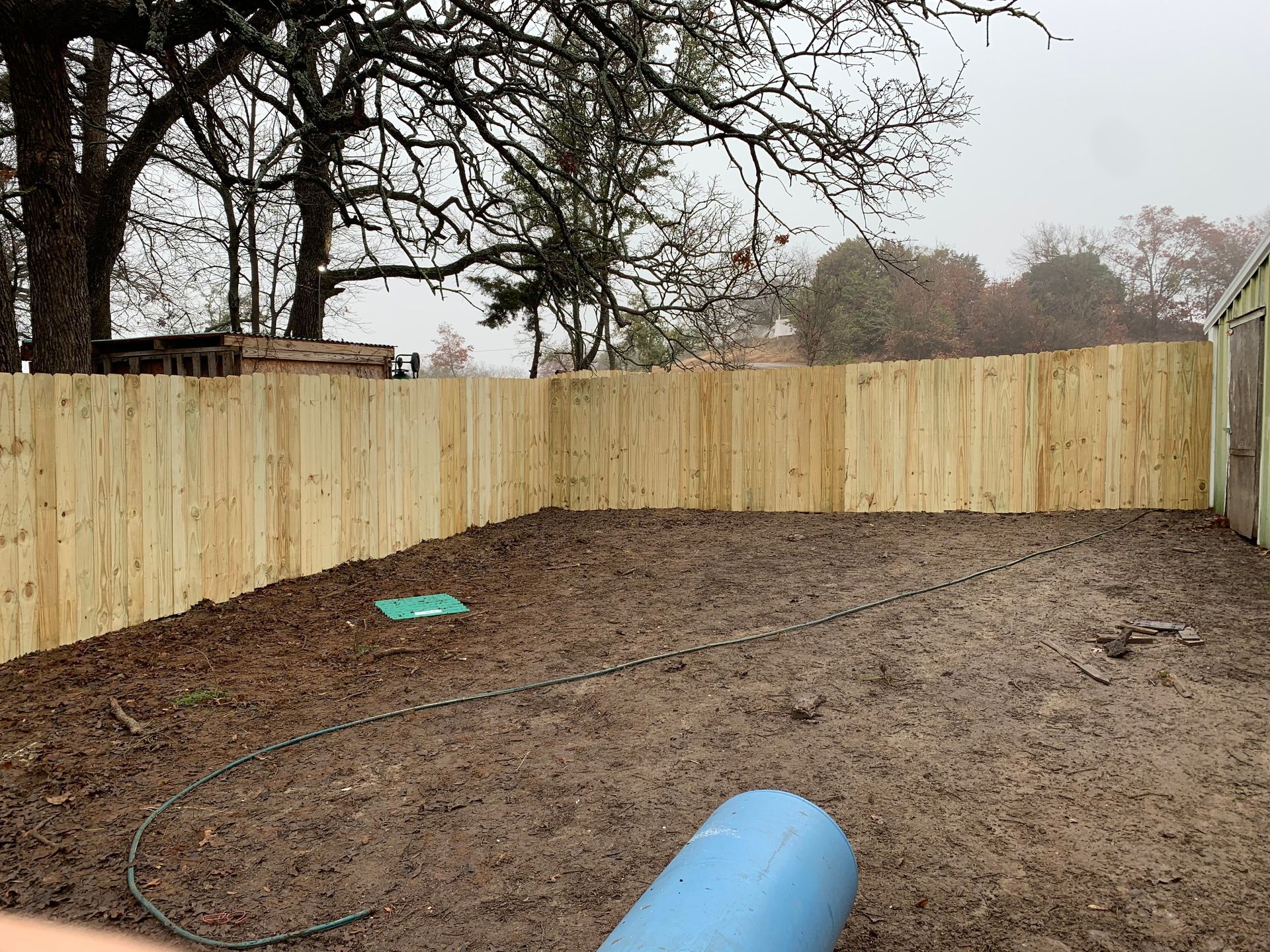 A newly installed wooden fence encloses a muddy, dirt-filled backyard with a blue barrel in the foreground.