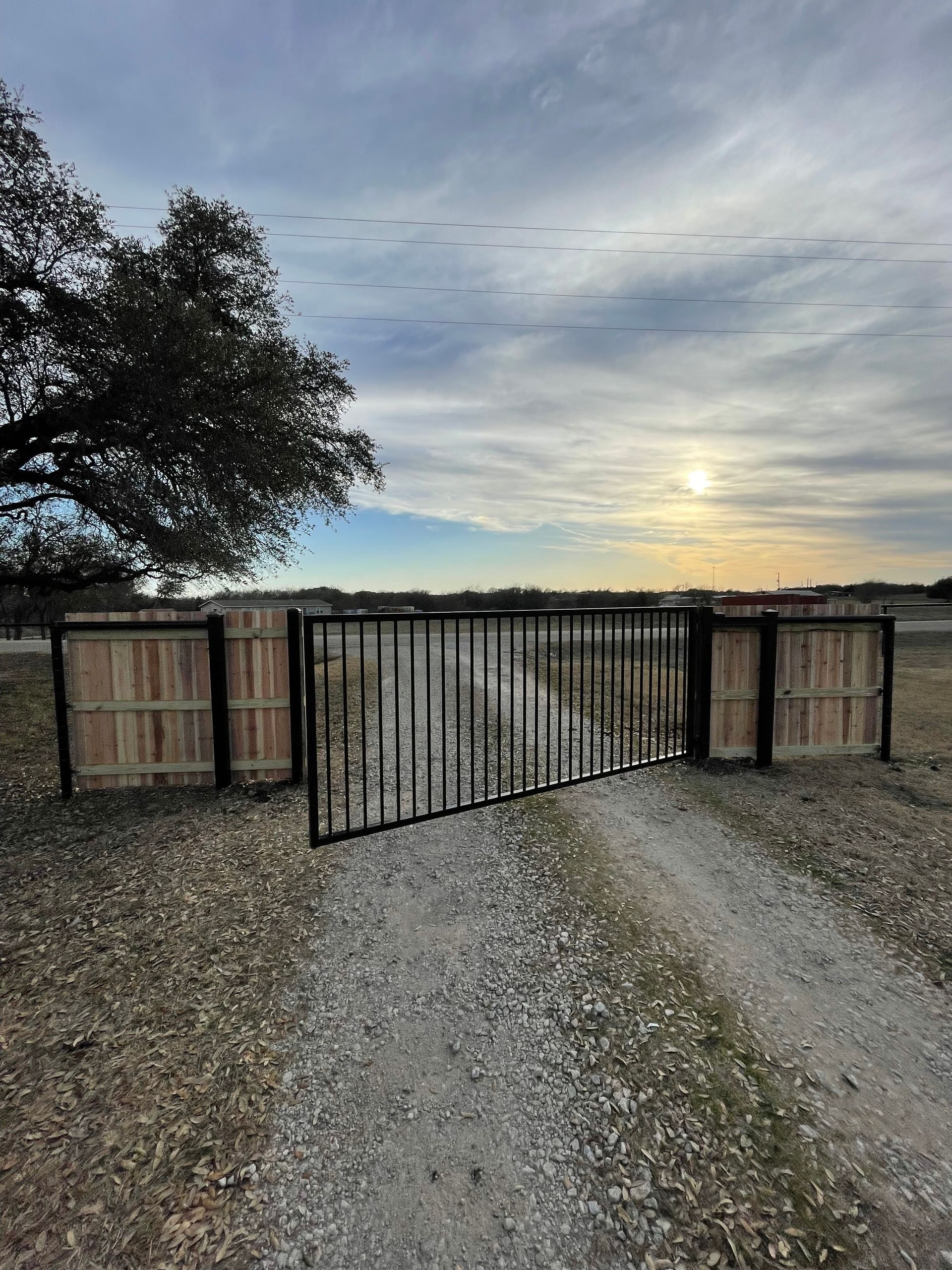 A black metal gate stands across a gravel driveway at sunset, flanked by wooden fence sections under a cloudy sky.