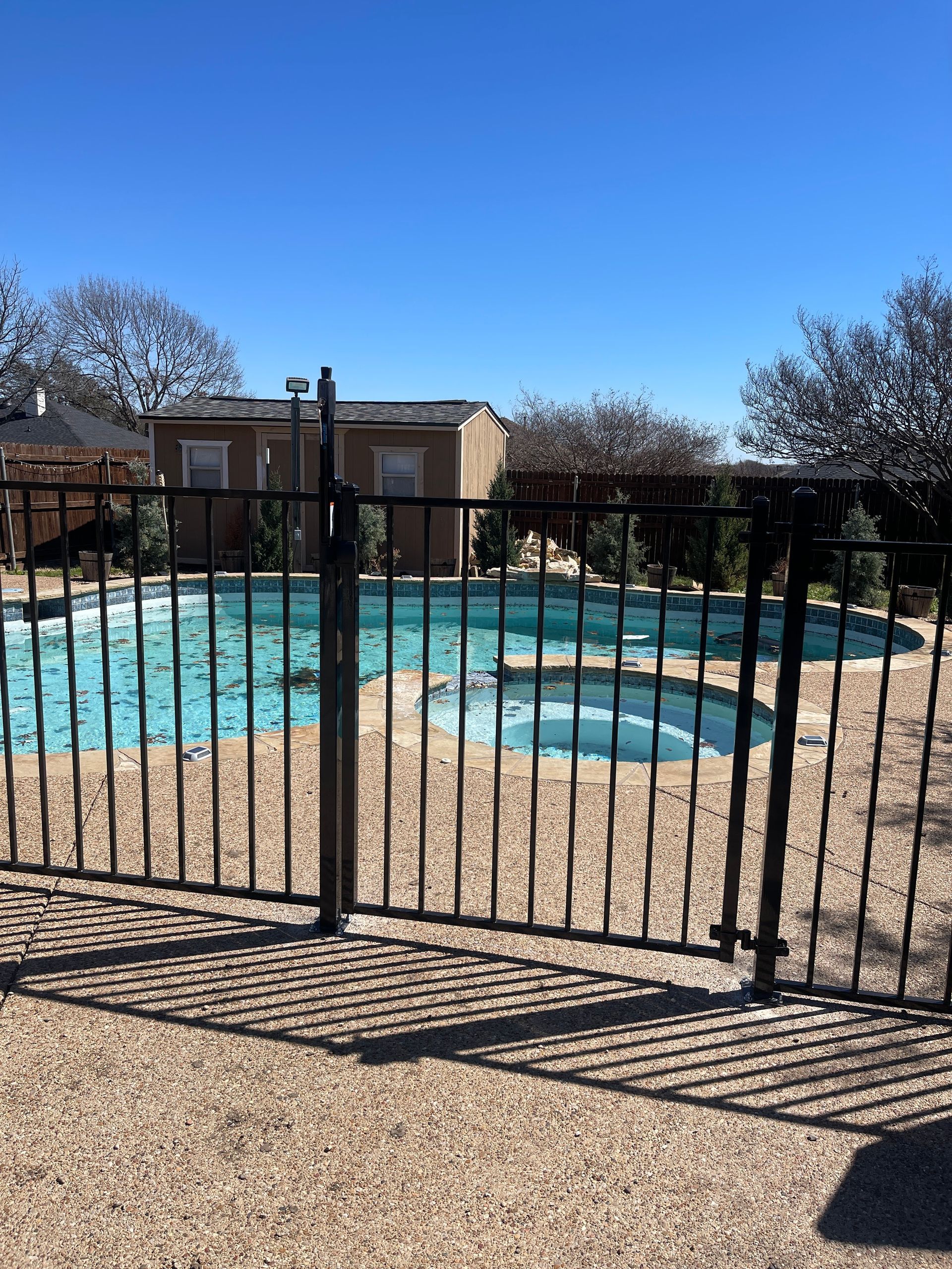 A black metal fence surrounds a backyard swimming pool and hot tub on a sunny day.
