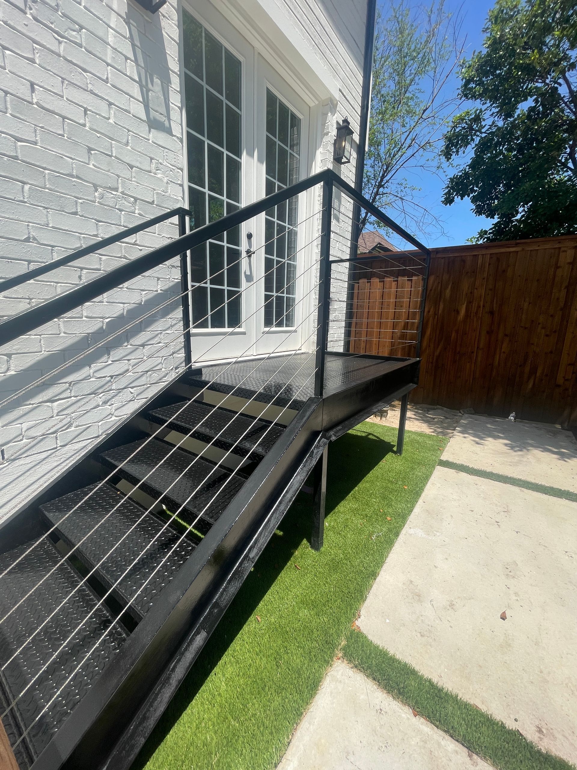 Black metal outdoor stairs with cable railings leading to white French doors against a brick wall and a grassy yard.