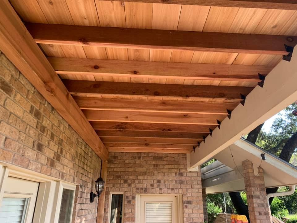 A covered outdoor patio featuring a natural wood-plank ceiling supported by wooden beams attached to a brick wall.