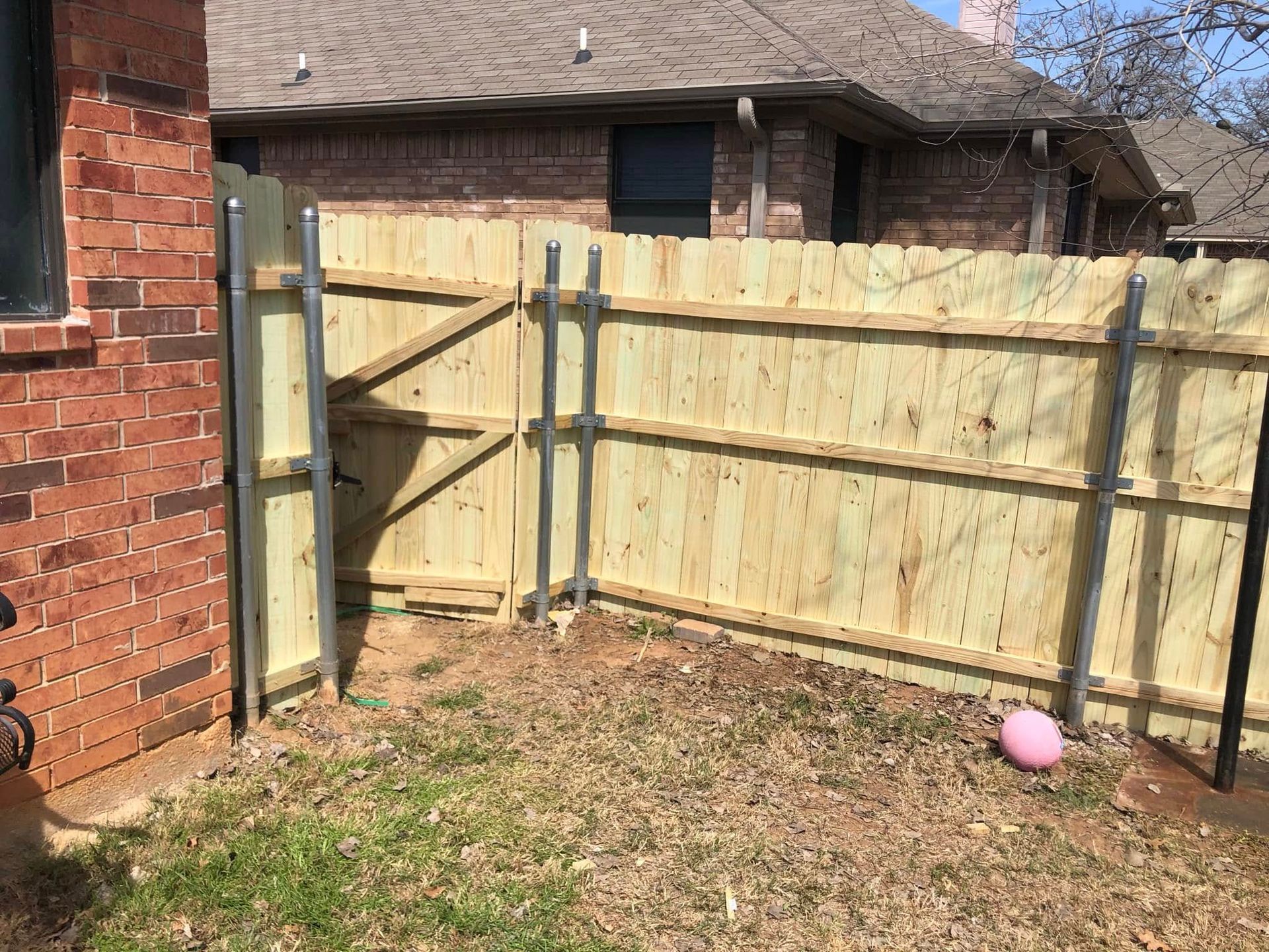 A new wooden privacy fence with metal posts stands in a backyard next to a brick house wall.