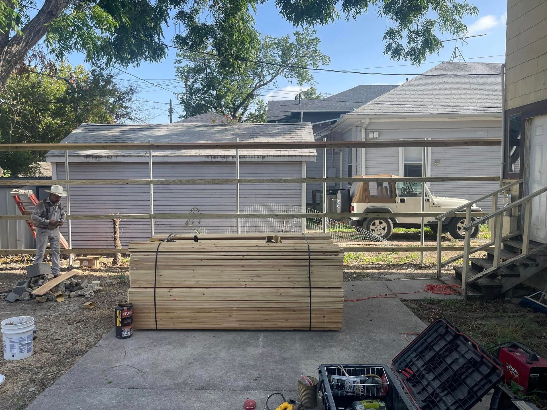 A construction worker stands near a large stack of lumber in a backyard with a metal fence and parked Jeep.