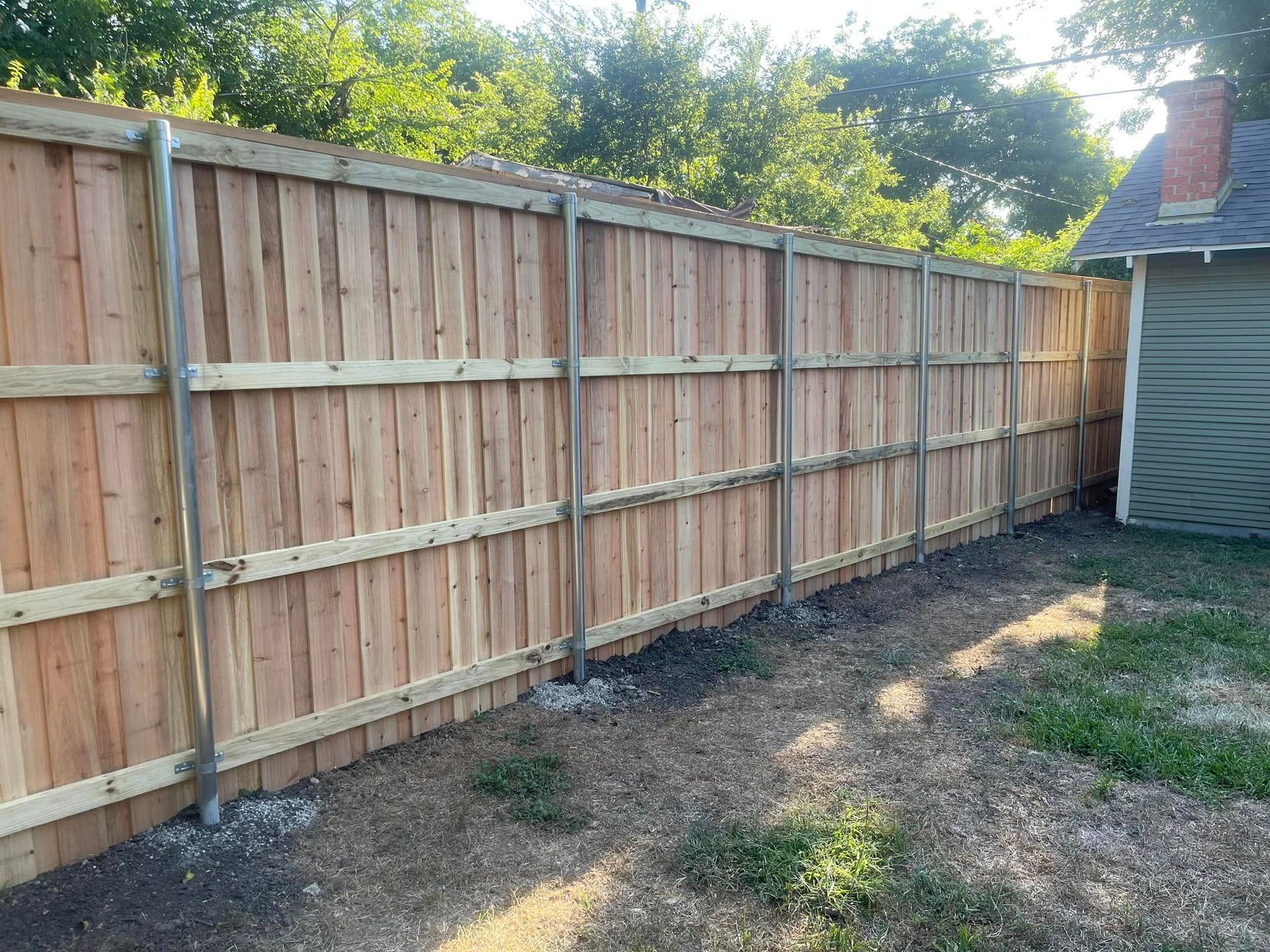 A newly installed wood privacy fence with metal support posts, set in a yard with a neighboring house visible.