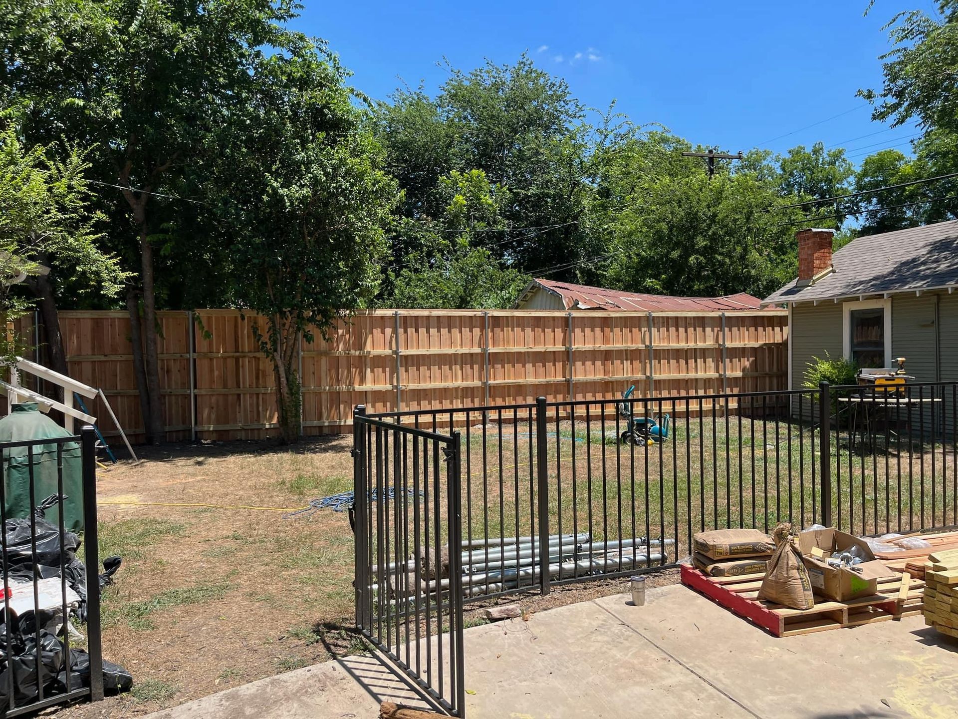 A backyard scene featuring a tall wooden privacy fence, a black metal gate, and construction materials on a concrete patio.