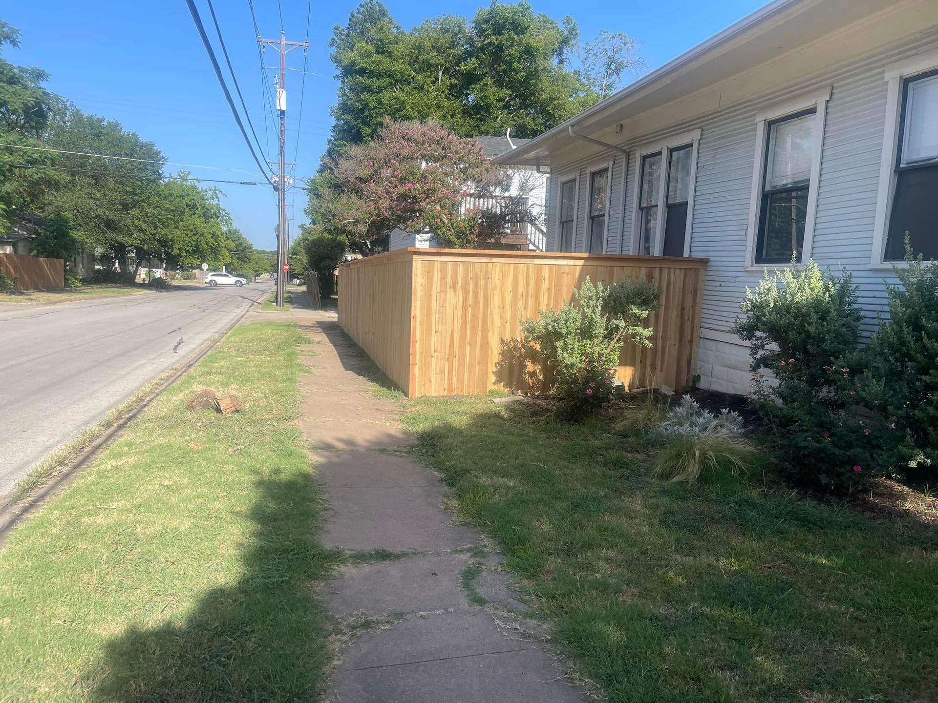 A wooden fence borders a sidewalk and a light-colored house on a sunny day next to a road with utility poles.
