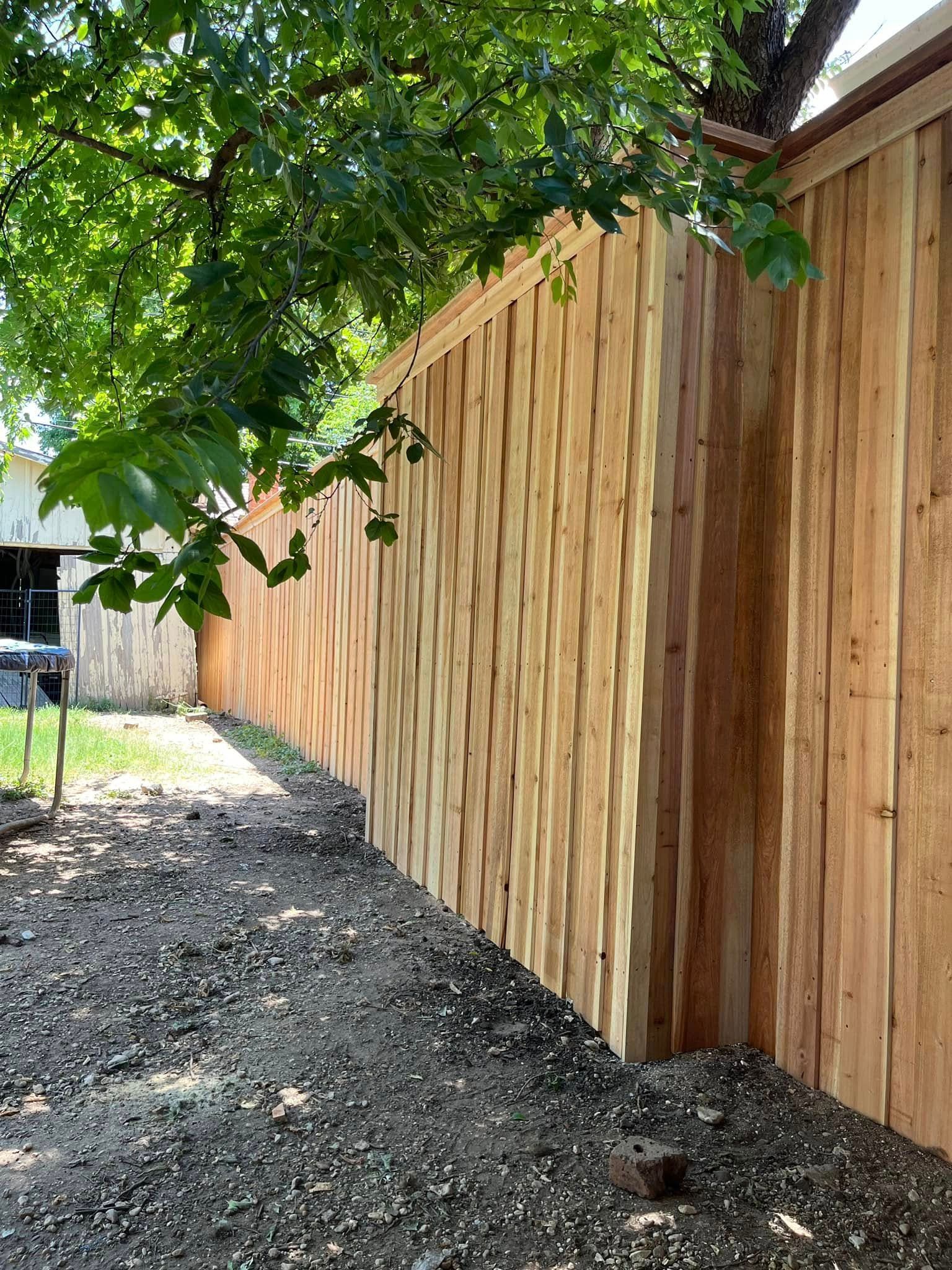 A new, light-brown wood fence made of vertical, rustic boards sits next to a dirt ground under a leafy green tree.