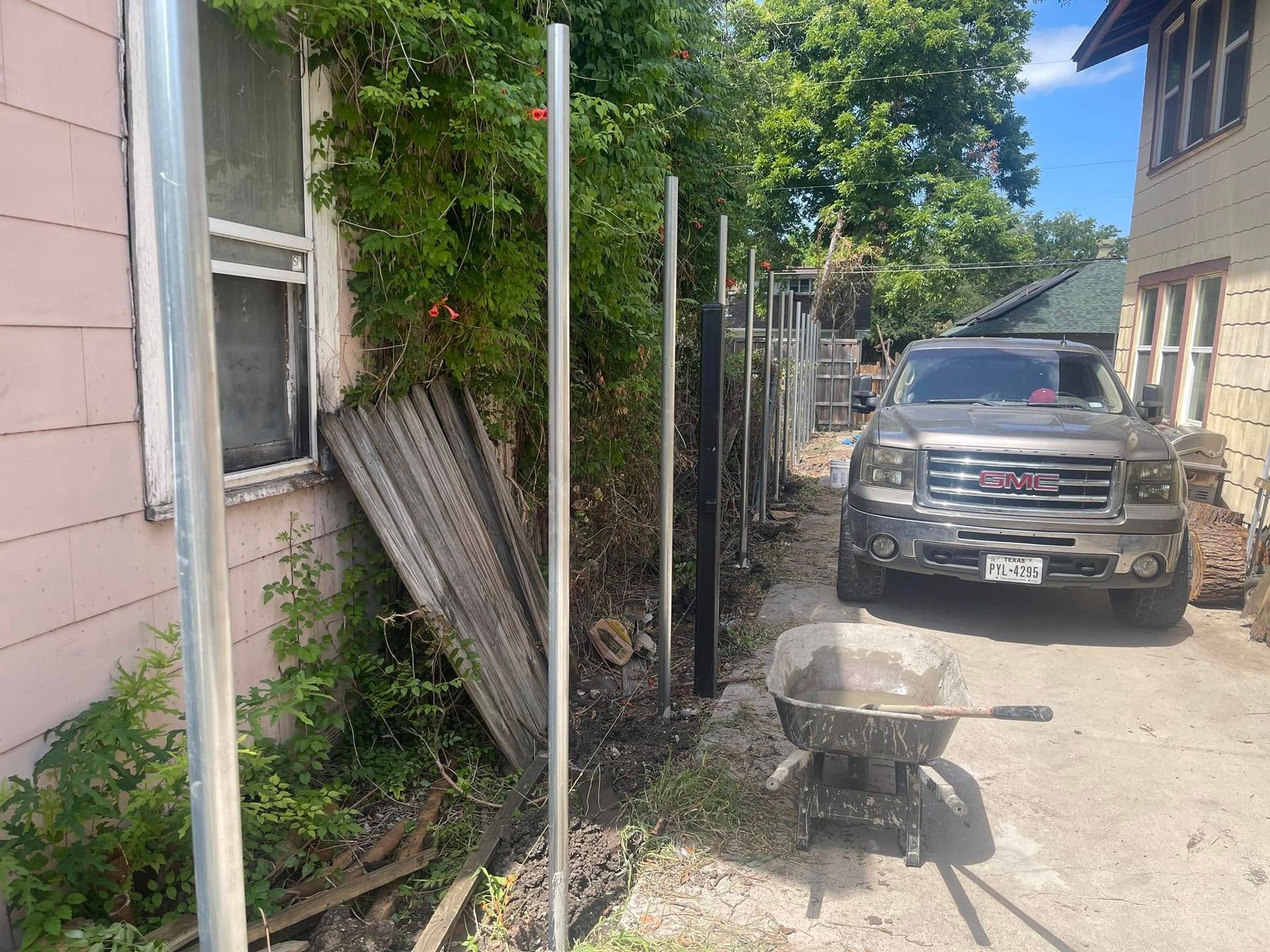 Metal fence posts stand in a line next to a house and driveway where a gray GMC truck is parked near a wheelbarrow.