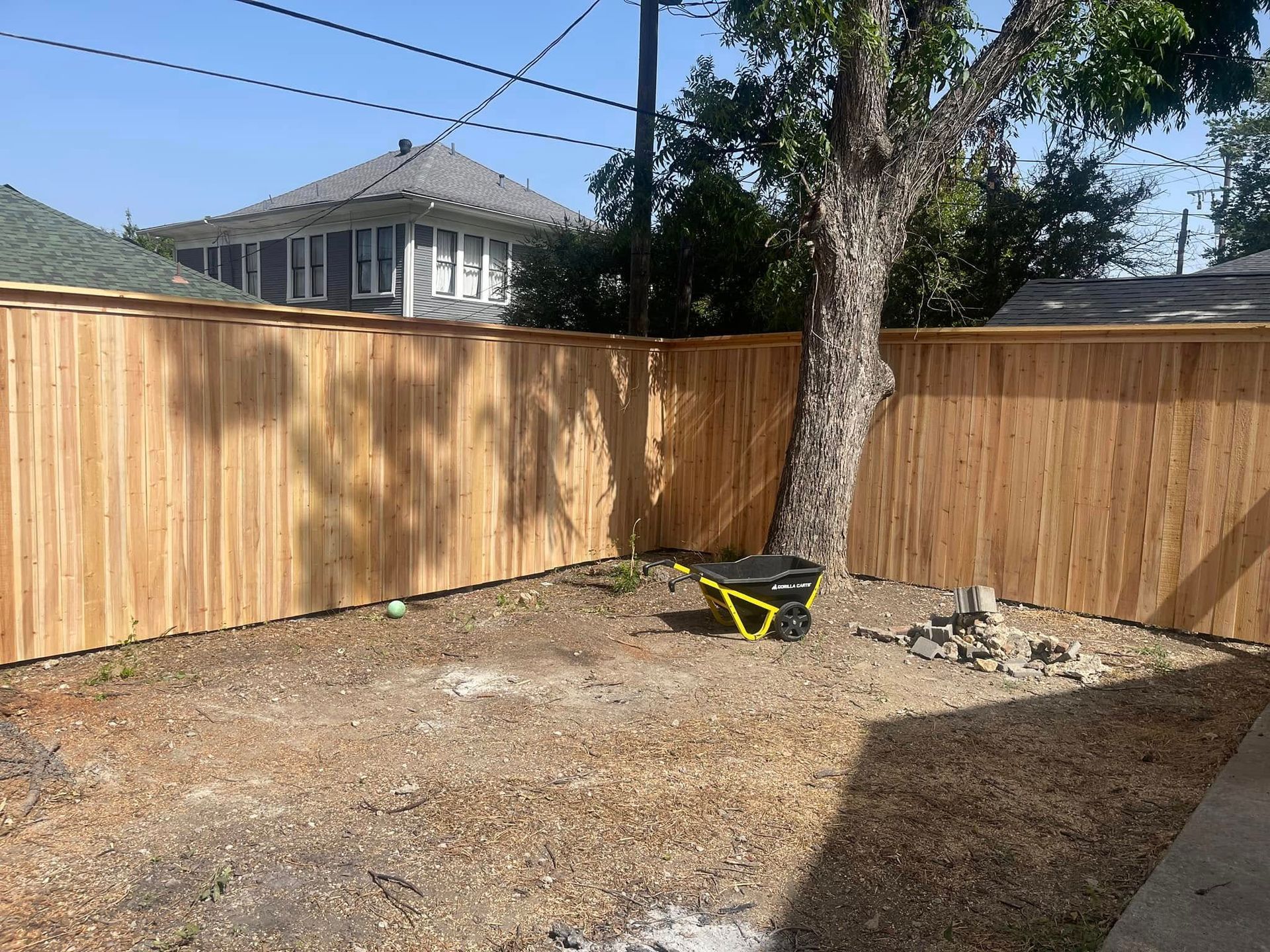 A newly built wooden privacy fence surrounds a backyard area with a large tree, gravel ground, and a small yellow cart.