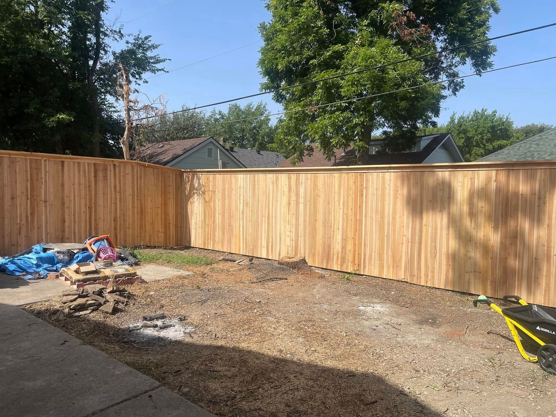 New tall wooden privacy fence in a backyard, with a patchy dirt yard, construction materials, and a yellow cart.