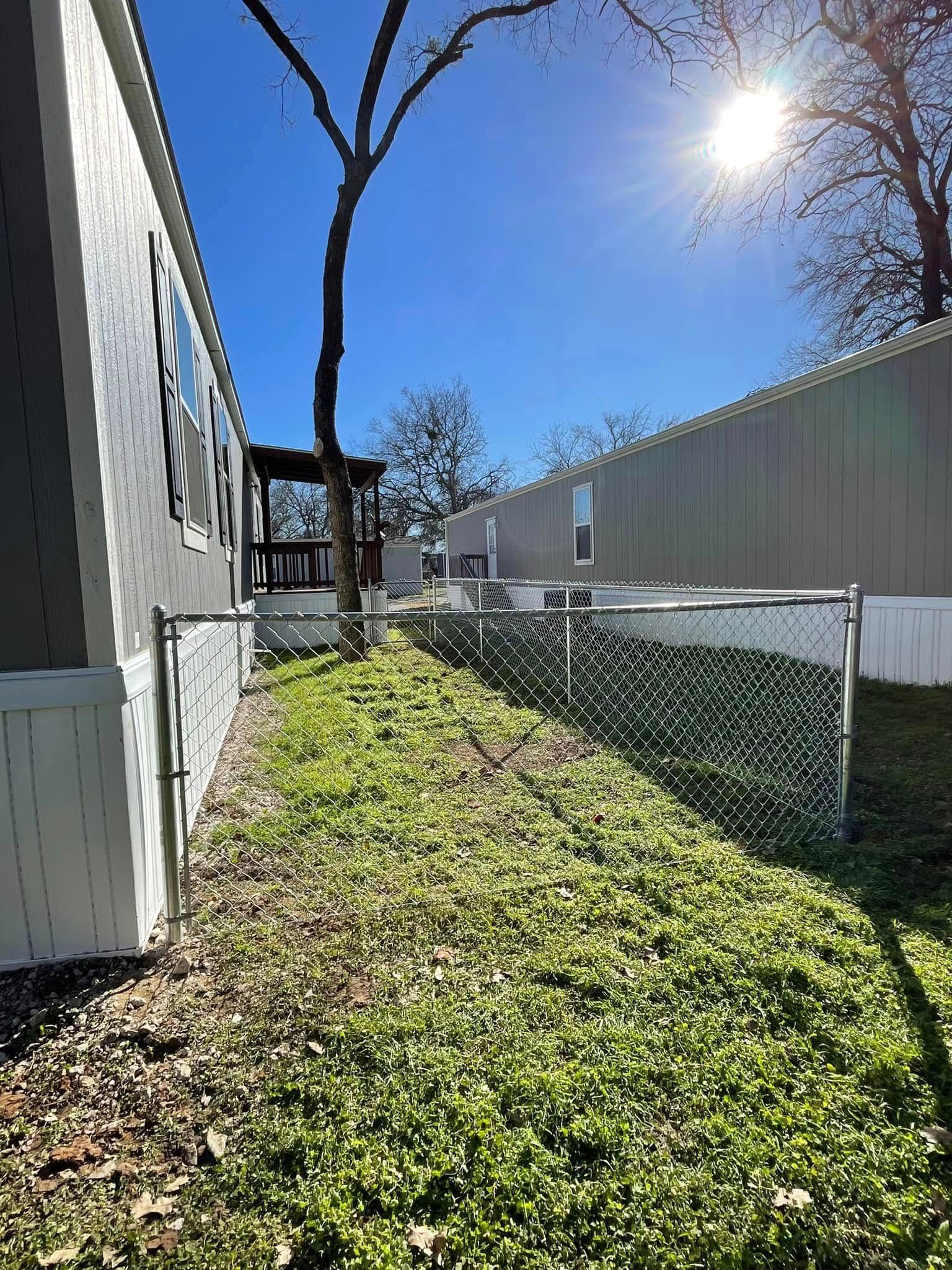 A chain-link fence divides a narrow, grassy yard between two light-colored mobile homes under a bright, sunny sky.