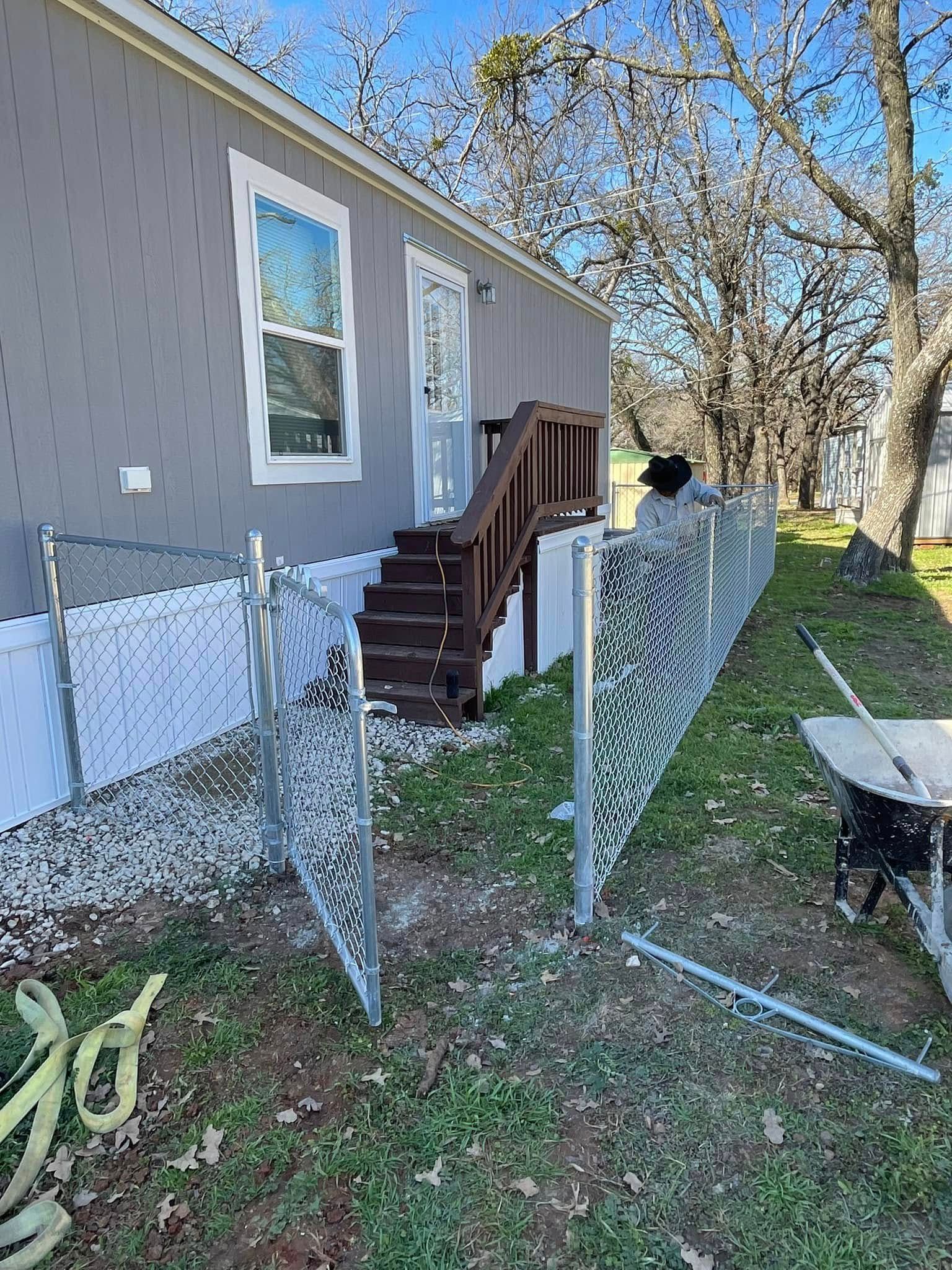 A person in a hat works on installing a chain-link fence beside the wooden stairs of a gray mobile home.