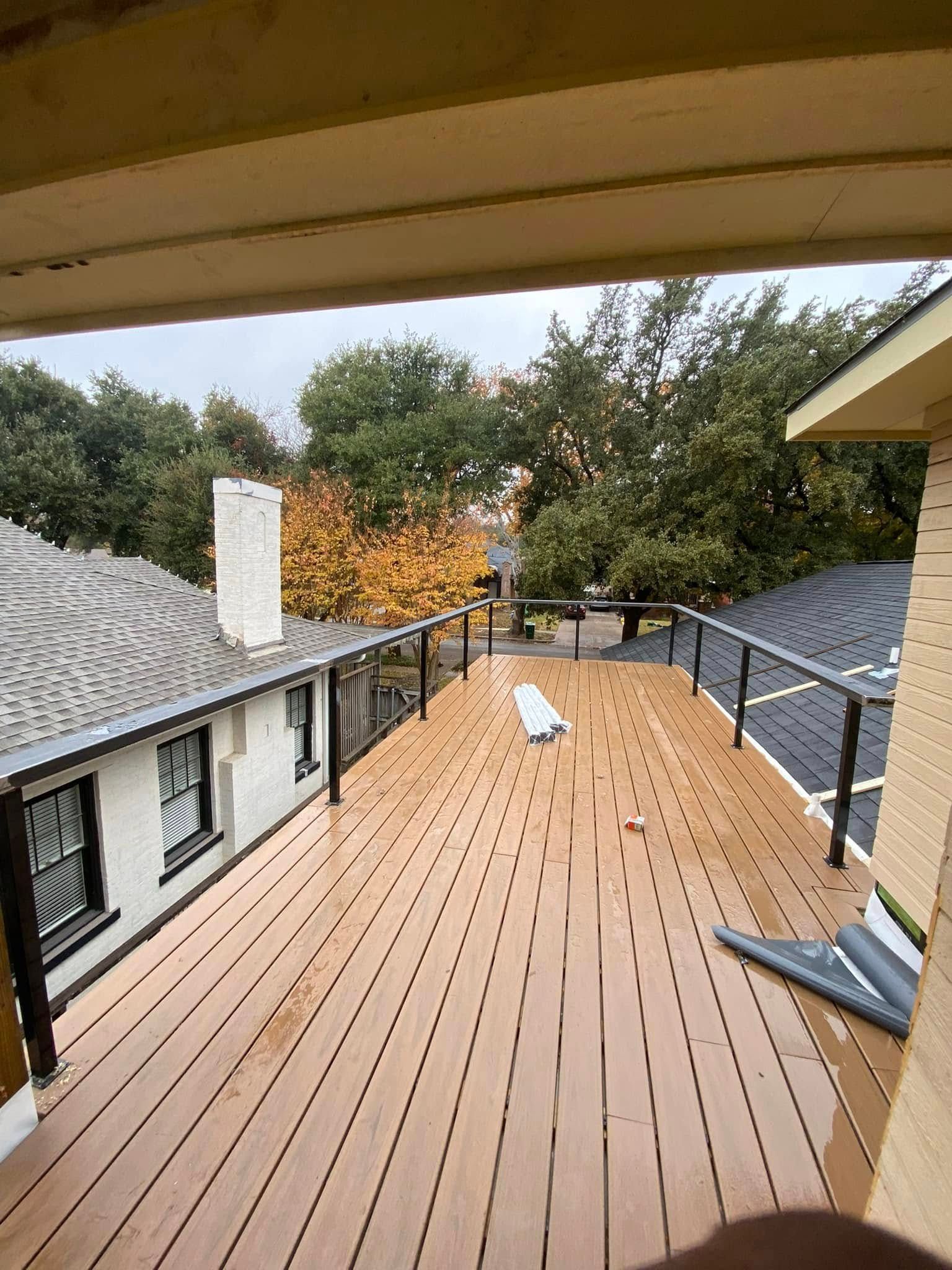 A newly installed light-brown wooden deck connects two rooftops with black railings under a cloudy sky with autumn trees.