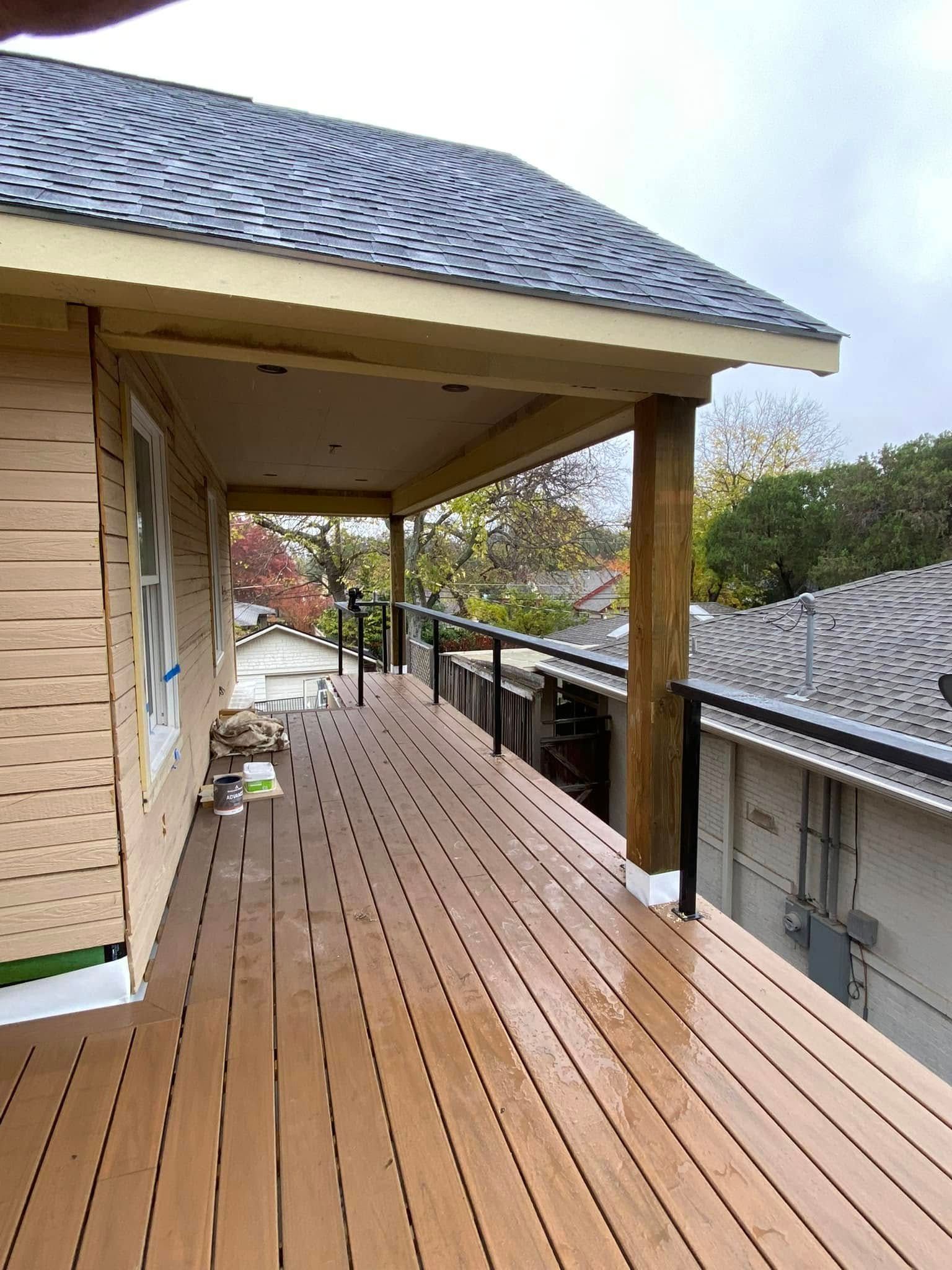 A wooden deck under a house roof overhang, featuring black railings and views of neighboring houses and trees.