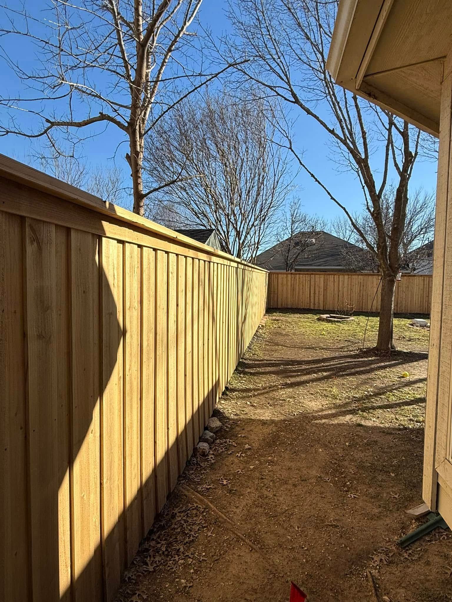 A wooden privacy fence lines a narrow yard leading to a back fence under a clear blue sky.