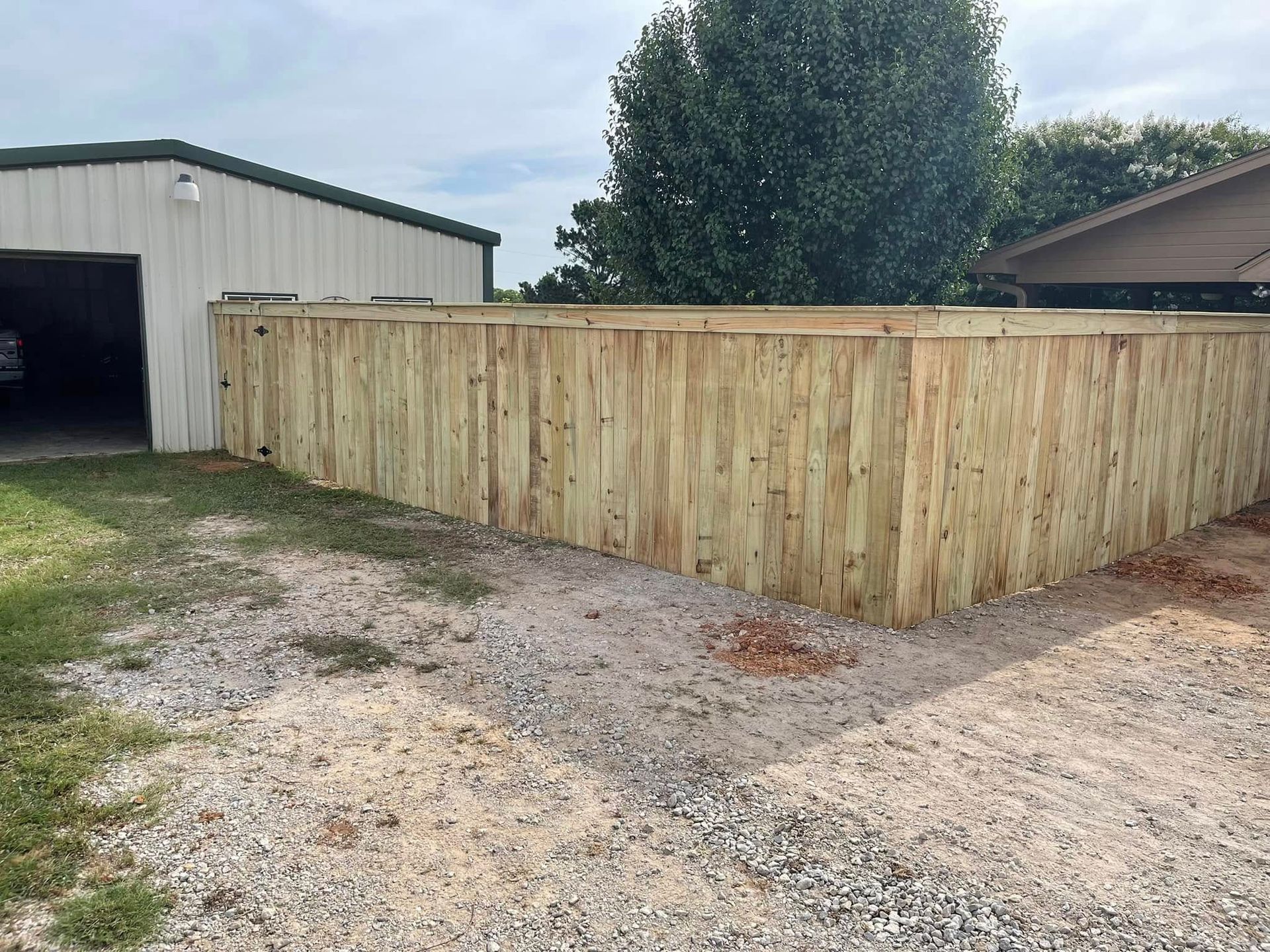 A new, light-colored wooden privacy fence stands in a gravel yard between a metal garage and a house.