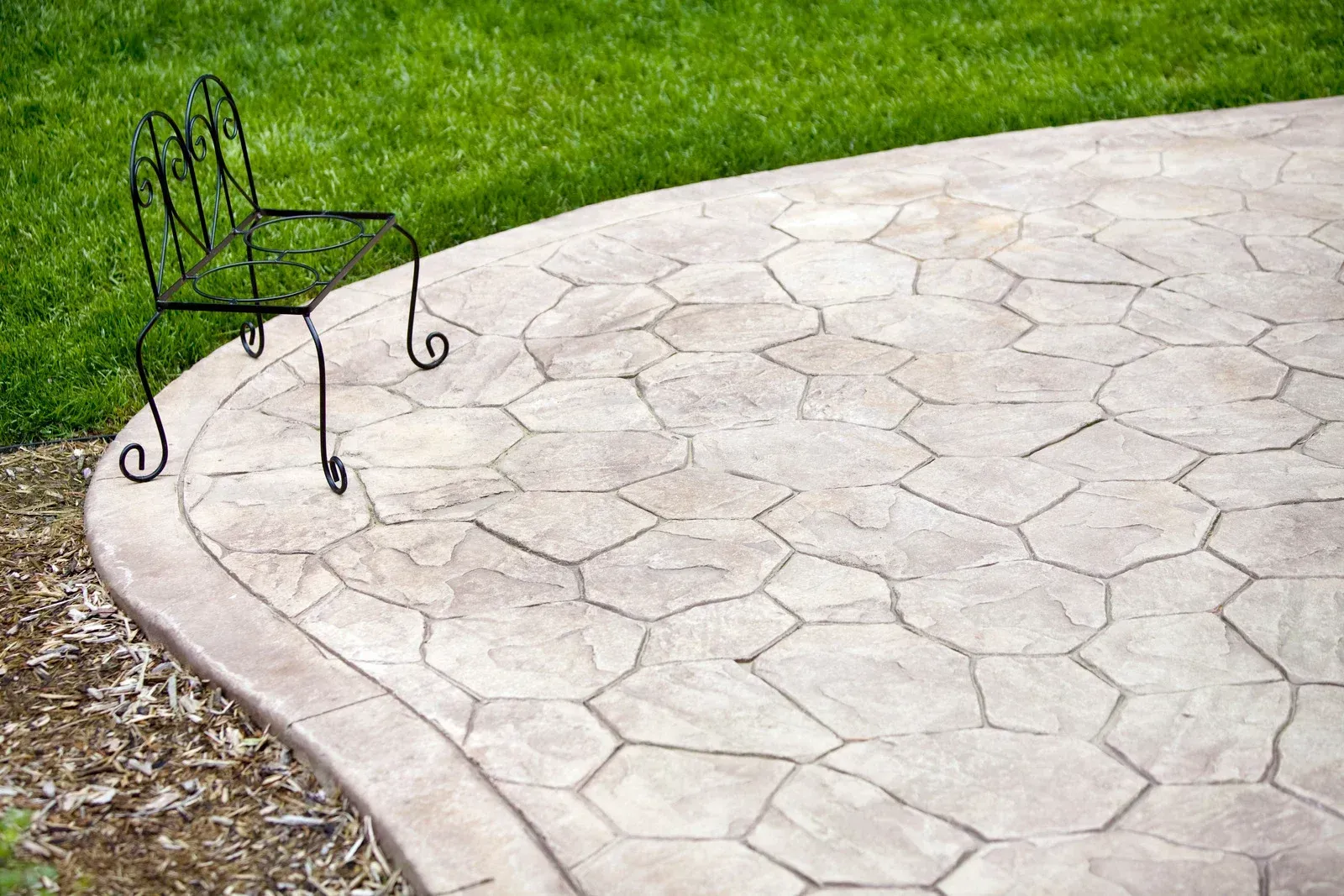 A small, ornate black metal chair sits on a curved, light-colored stamped concrete patio next to a green grassy lawn.