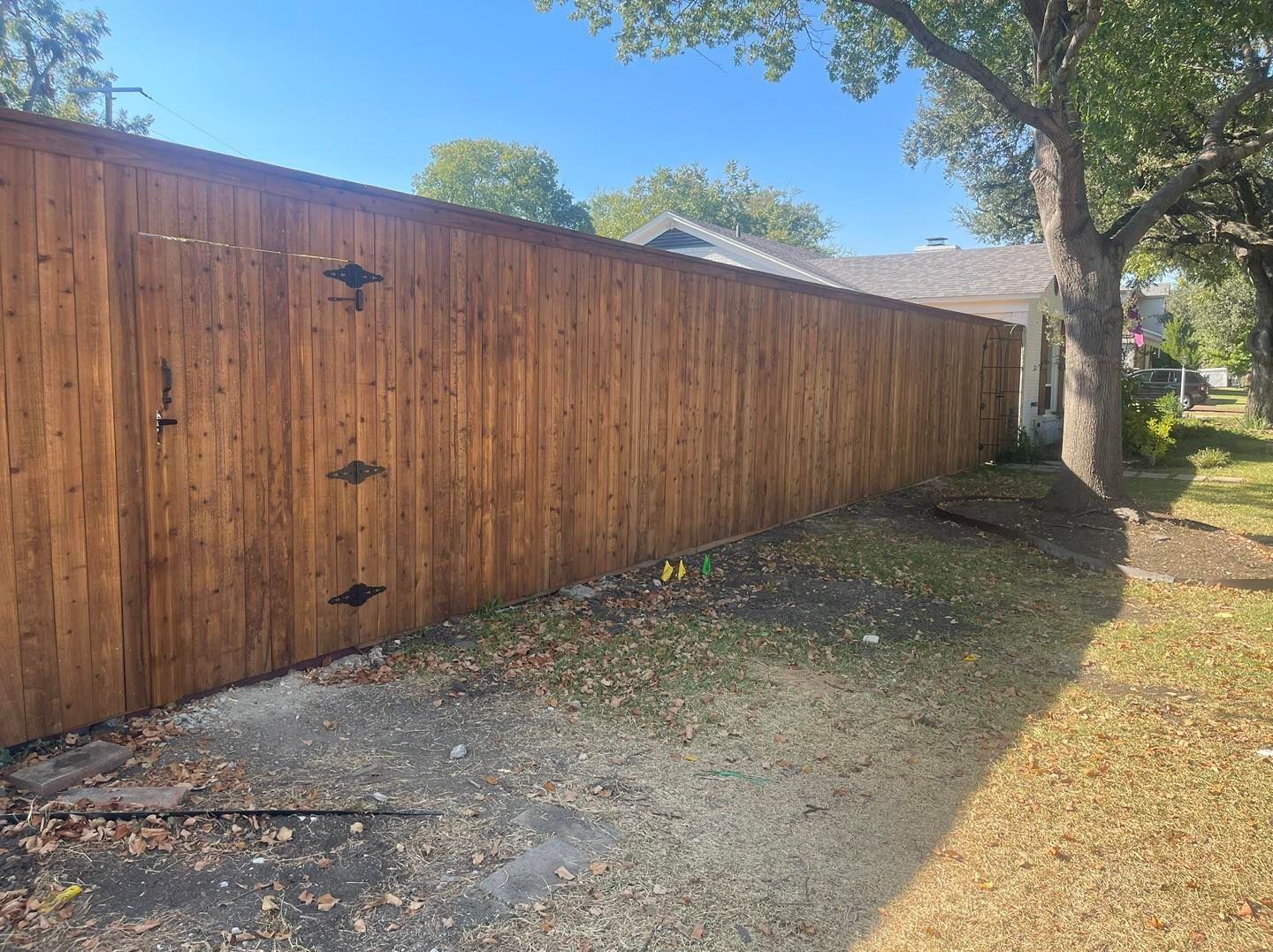 A long, stained wood privacy fence with a gated section, situated on a grassy lot next to a house and a large tree.