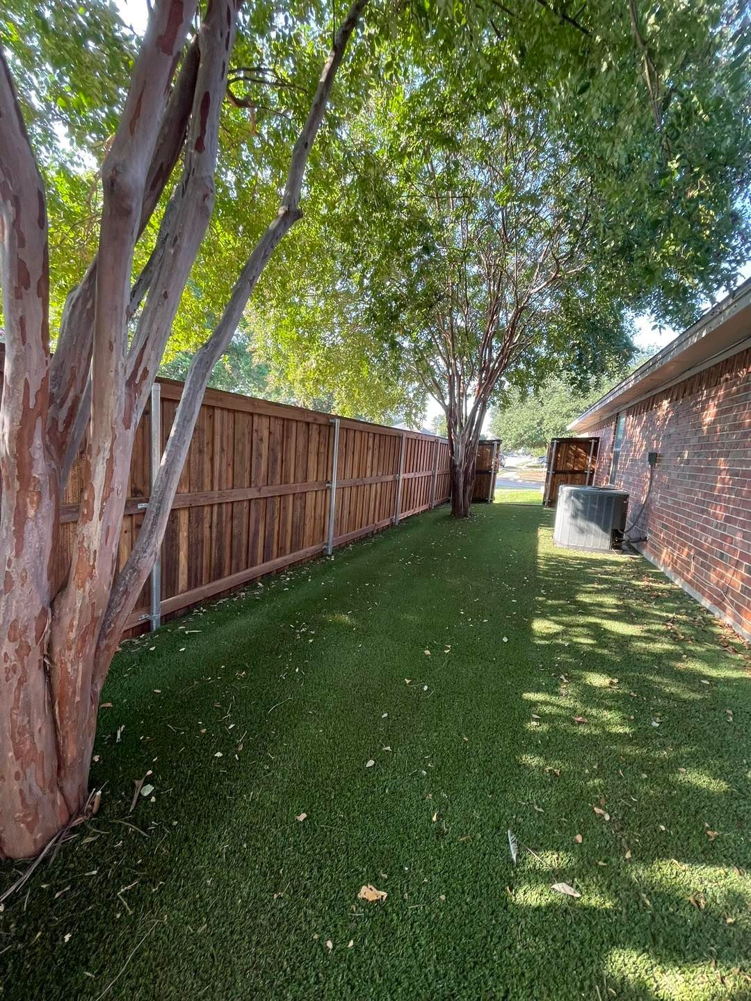 A narrow side yard with a wooden fence, green lawn, and trees alongside a brick house exterior.