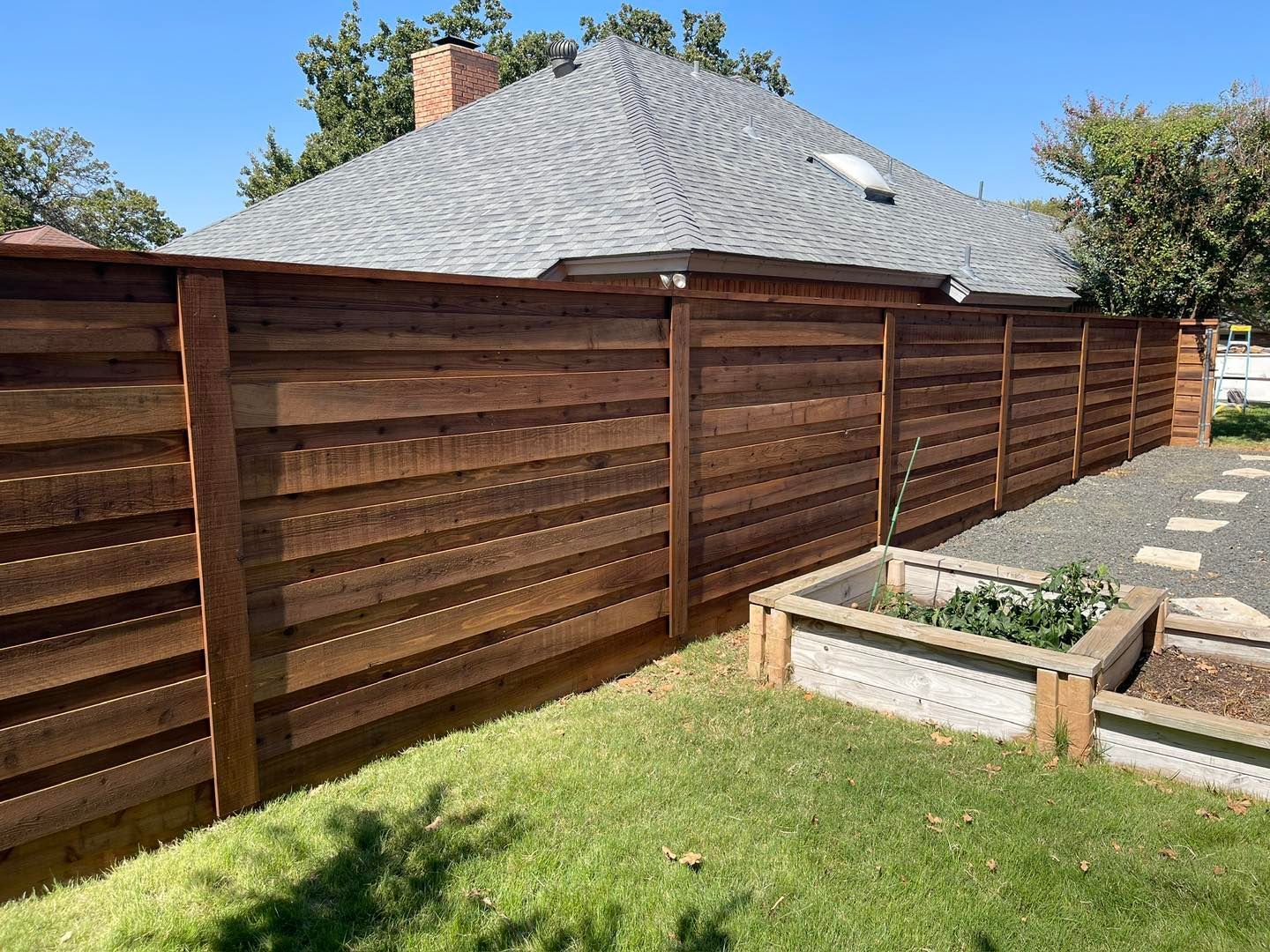 A modern horizontal cedar privacy fence runs along a backyard next to a residential house and stone garden beds.