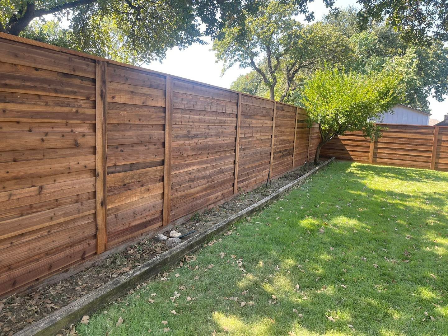 A long, horizontal wooden fence with a stained brown finish sits above a stone border next to a green lawn.