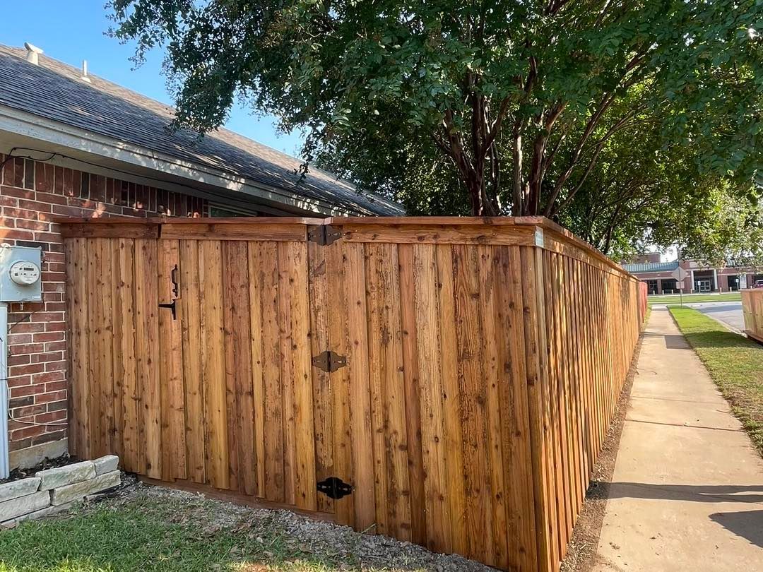 A new cedar wood fence stands beside a brick house and a public concrete sidewalk under a clear blue sky.