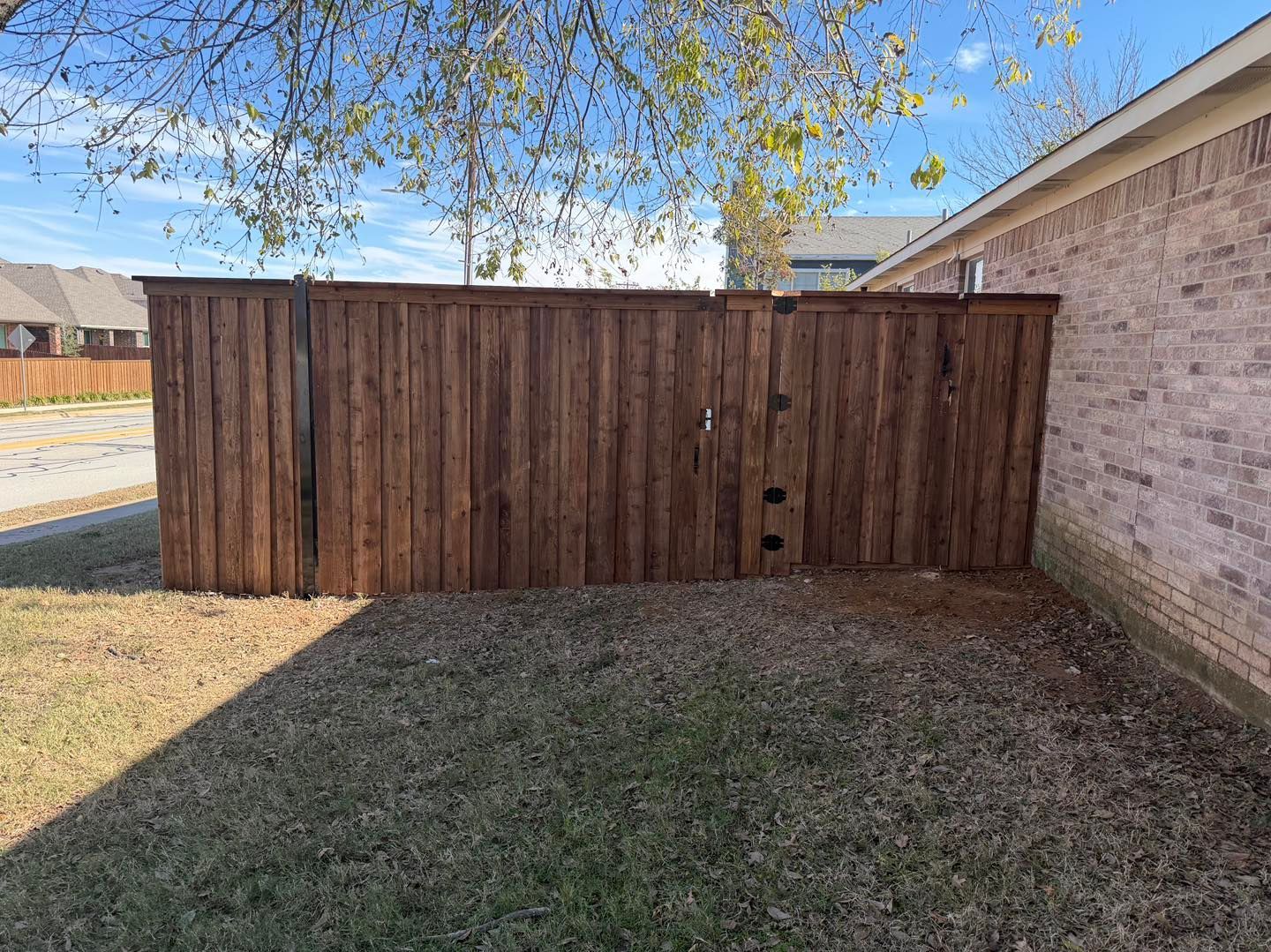 A wooden privacy fence extends from a brick house wall across a patch of grass on a sunny day.