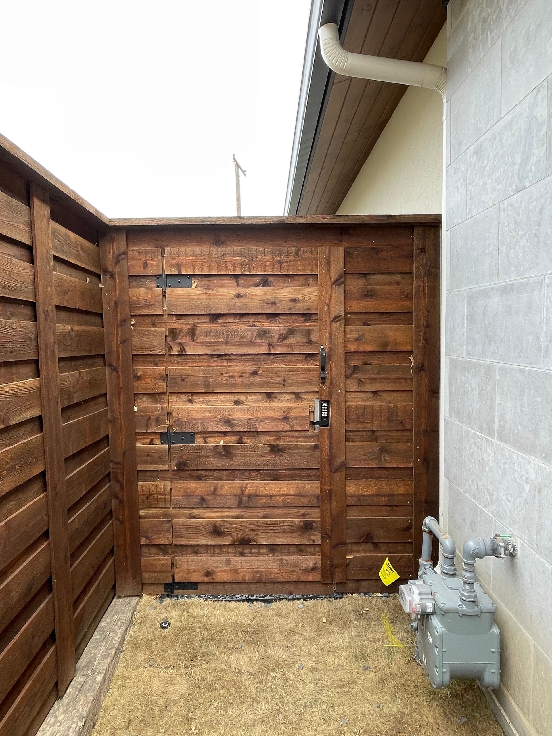 A wooden fence and gate in a side yard next to a stucco house wall with an exterior gas meter.