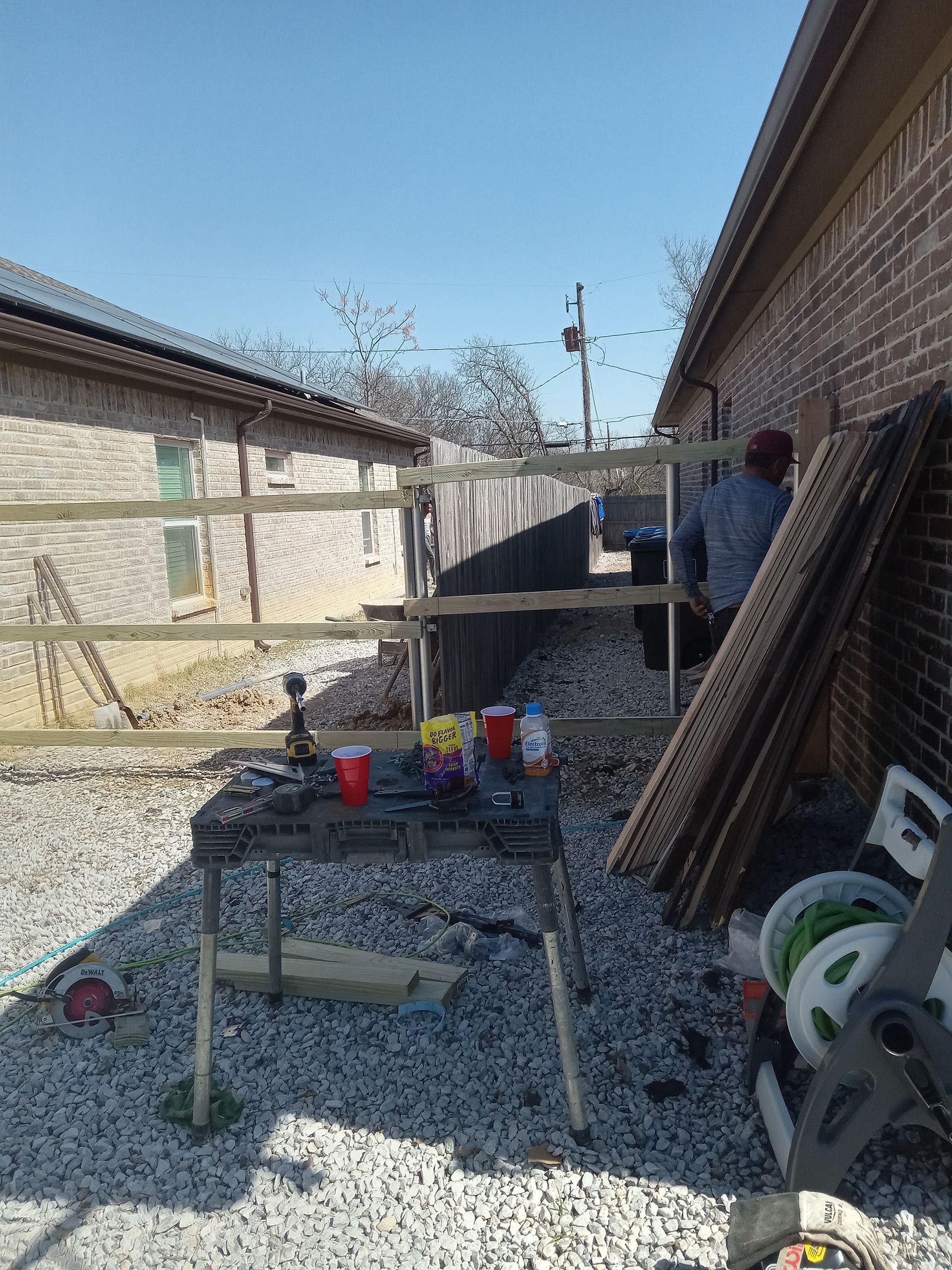 A person stands near a wooden fence being constructed between two brick houses on a gravel ground with a work table.