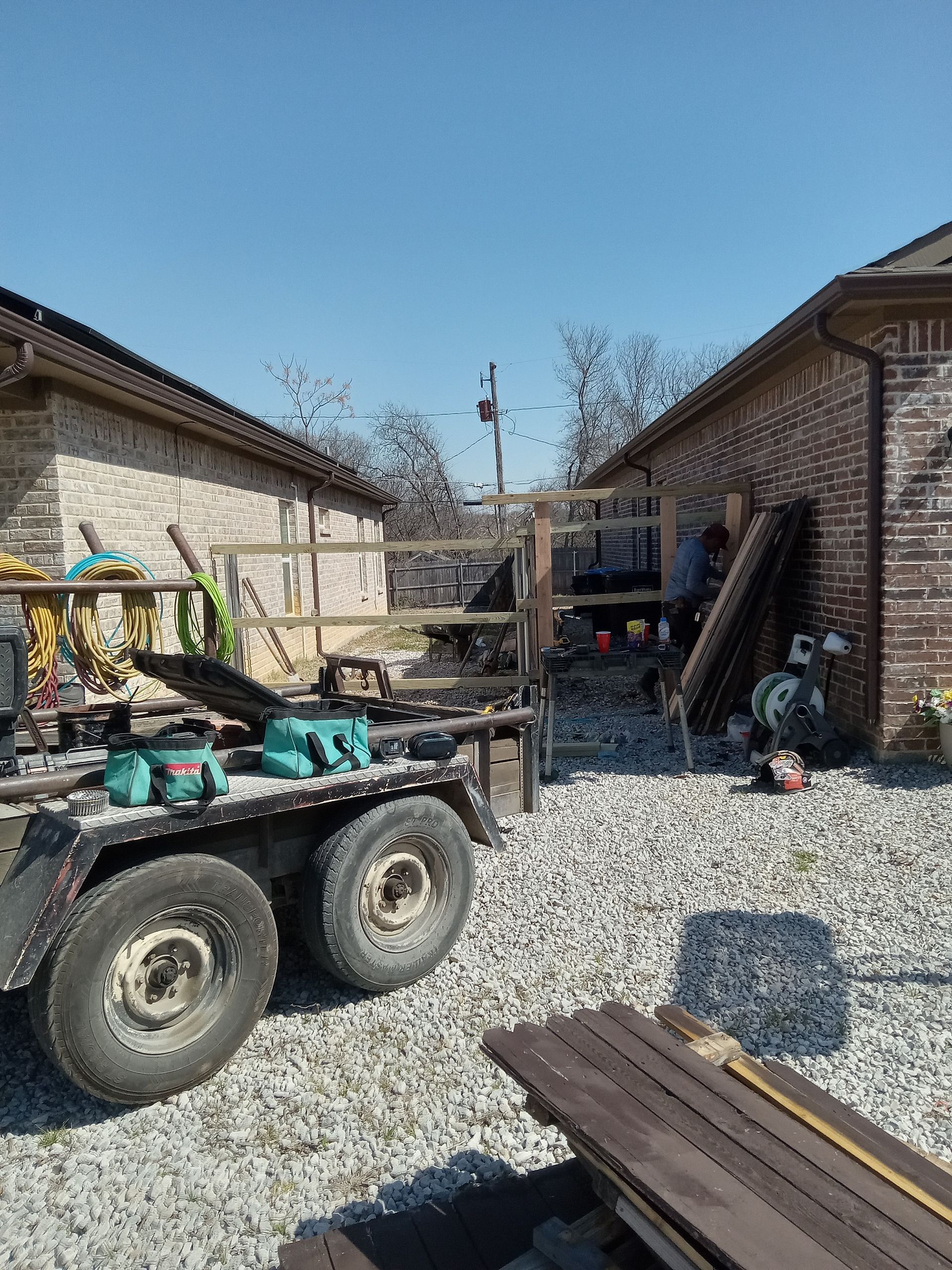 Utility trailer with equipment parked on gravel between two brick houses under a clear blue sky.