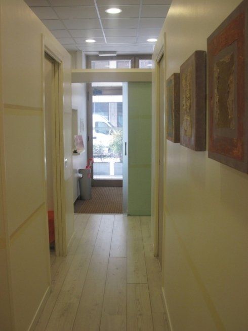 Stretto corridoio d'ufficio con pavimento in legno chiaro, quadri incorniciati e una porta a vetri in fondo.