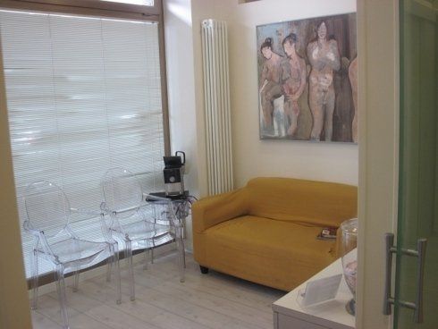 Piccola sala d'attesa con sedie trasparenti, divano color senape, tende e quadri alle pareti.
