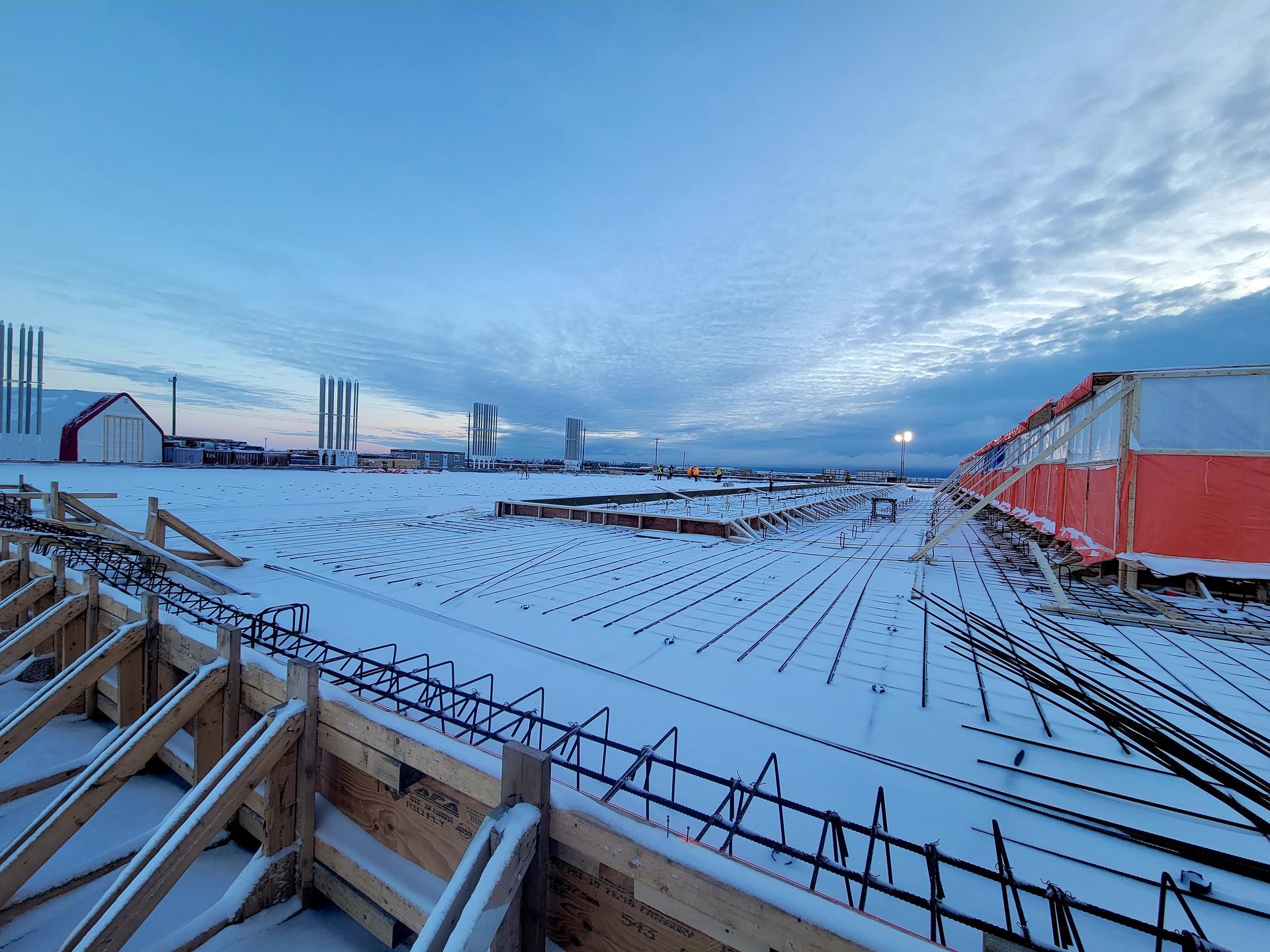 Chantier de construction enneigé en plein air, avec des poutres d'acier et des bâtiments rouges sous un ciel bleu nuageux au crépuscule