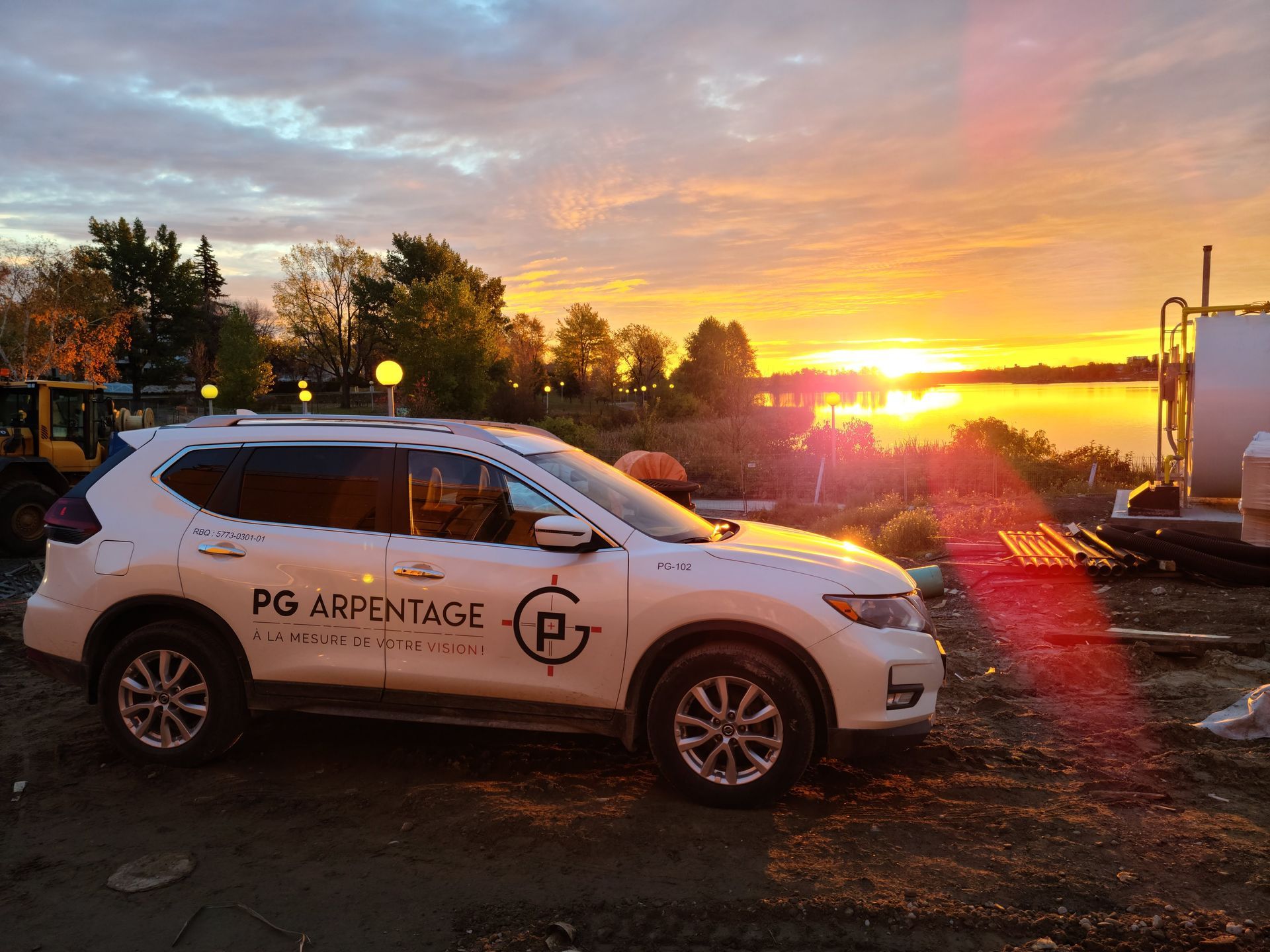 SUV blanc PG Arpentage stationné au lever du soleil sur un terrain gravillonné, avec des arbres et du matériel en arrière-plan.