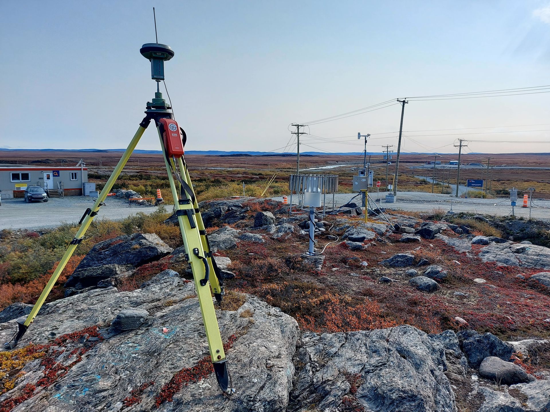 Un trépied d'arpenteur jaune équipé d'un récepteur GPS se dresse sur un sol rocailleux, semblable à la toundra, près d'une station de surveillance météorologique.
