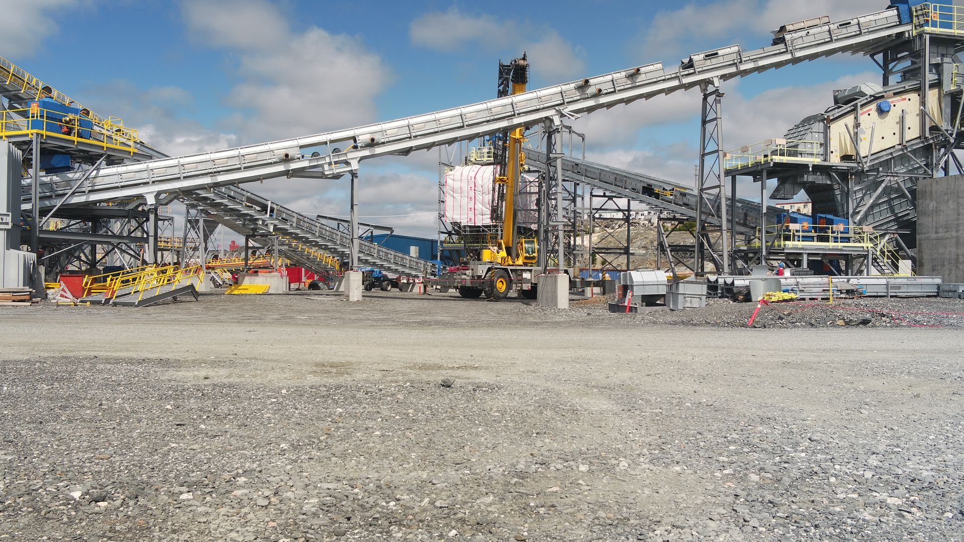 Site industriel avec de grands convoyeurs, des machines lourdes et des structures métalliques sous un ciel partiellement nuageux.
