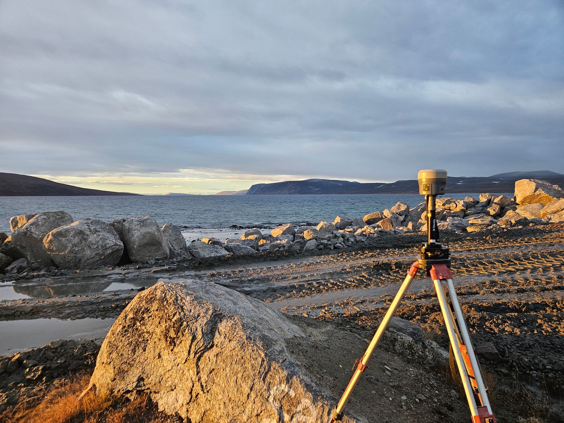 Station de mesure GPS sur trépied face à une côte rocheuse avec baie et montagnes en arrière-plan.