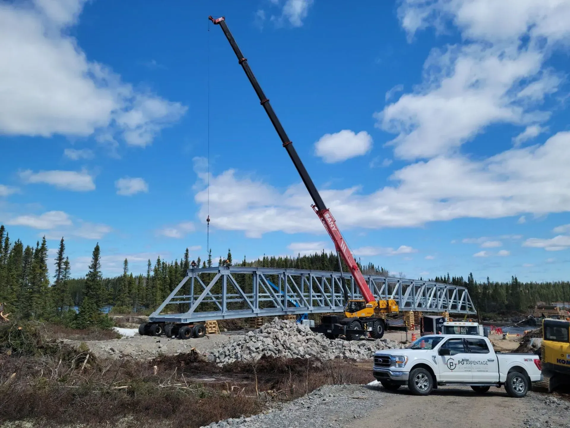 Une grue soulève une poutre métallique de pont au-dessus d'un chantier de construction, sous un ciel bleu et des camions.