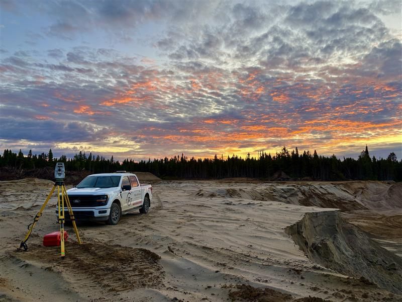 Un SUV blanc garé sur un chantier sablonneux au coucher du soleil, sous un ciel nuageux.
