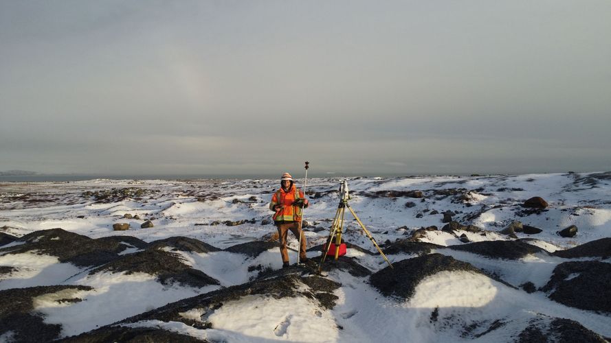 Une personne vêtue d'une tenue haute visibilité inspecte un paysage volcanique enneigé et rocailleux à l'aide d'équipement topographique professionnel.