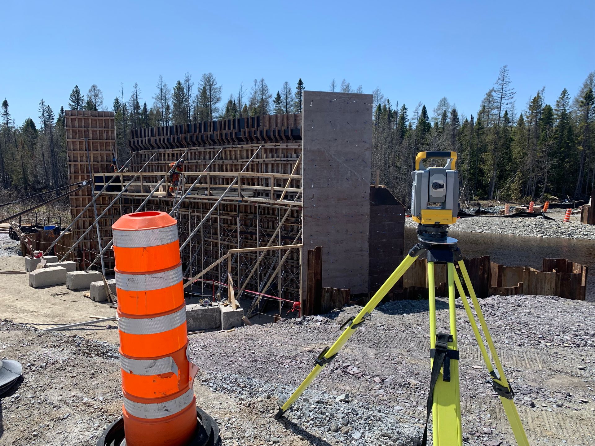 Chantier de construction avec équipement d'arpentage sur trépied jaune-vert, cône de signalisation et structures métalliques.