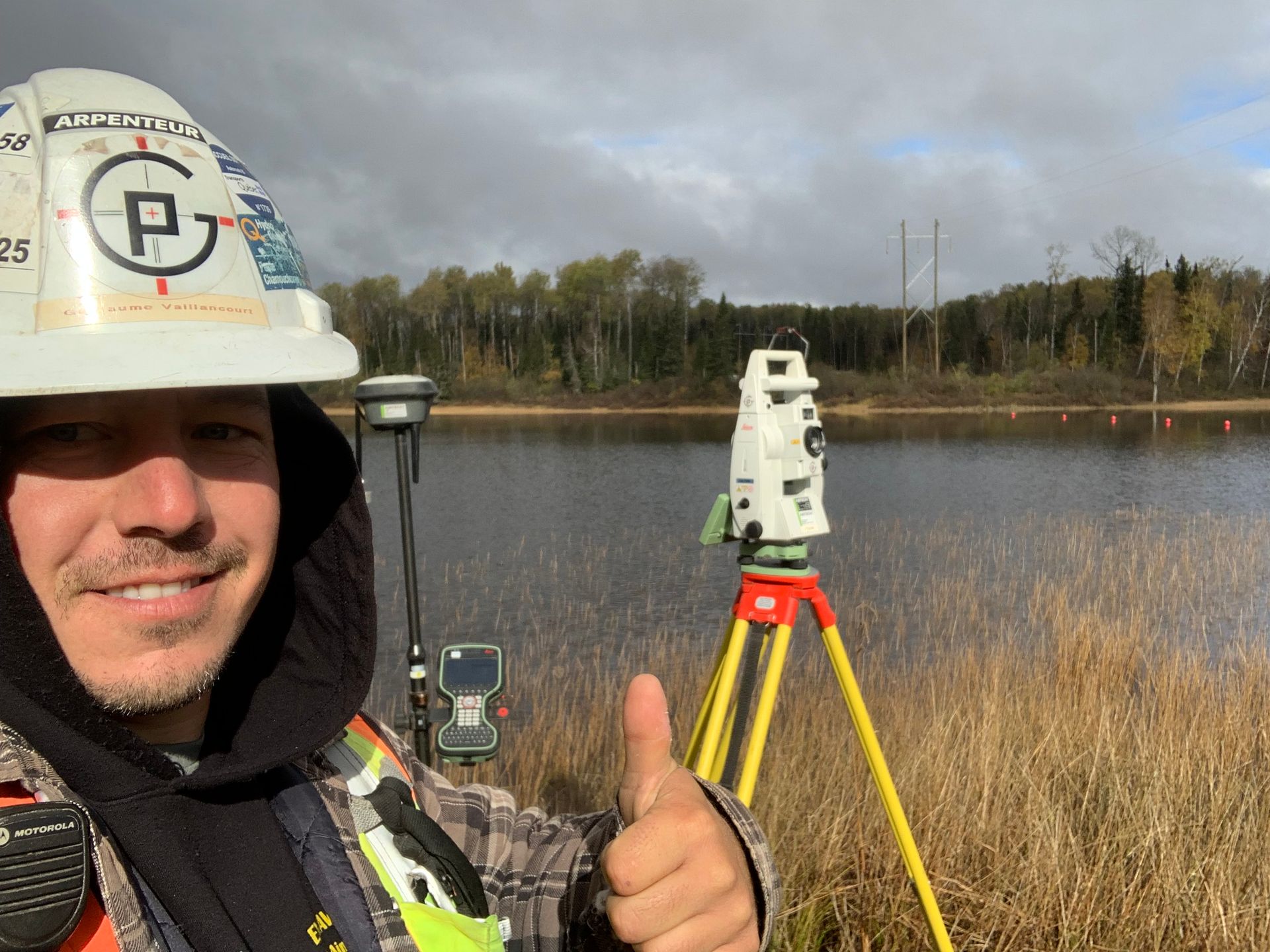Un géomètre portant un casque de chantier lève le pouce à côté d'un appareil topographique installé sur un trépied, avec un lac et des arbres en arrière-plan.