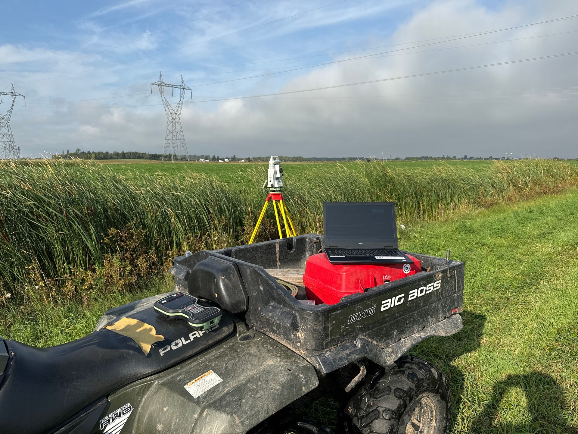 Une station totale de topographie sur trépied dans un champ herbeux, à côté d'un quad avec un ordinateur portable sur la benne arrière.